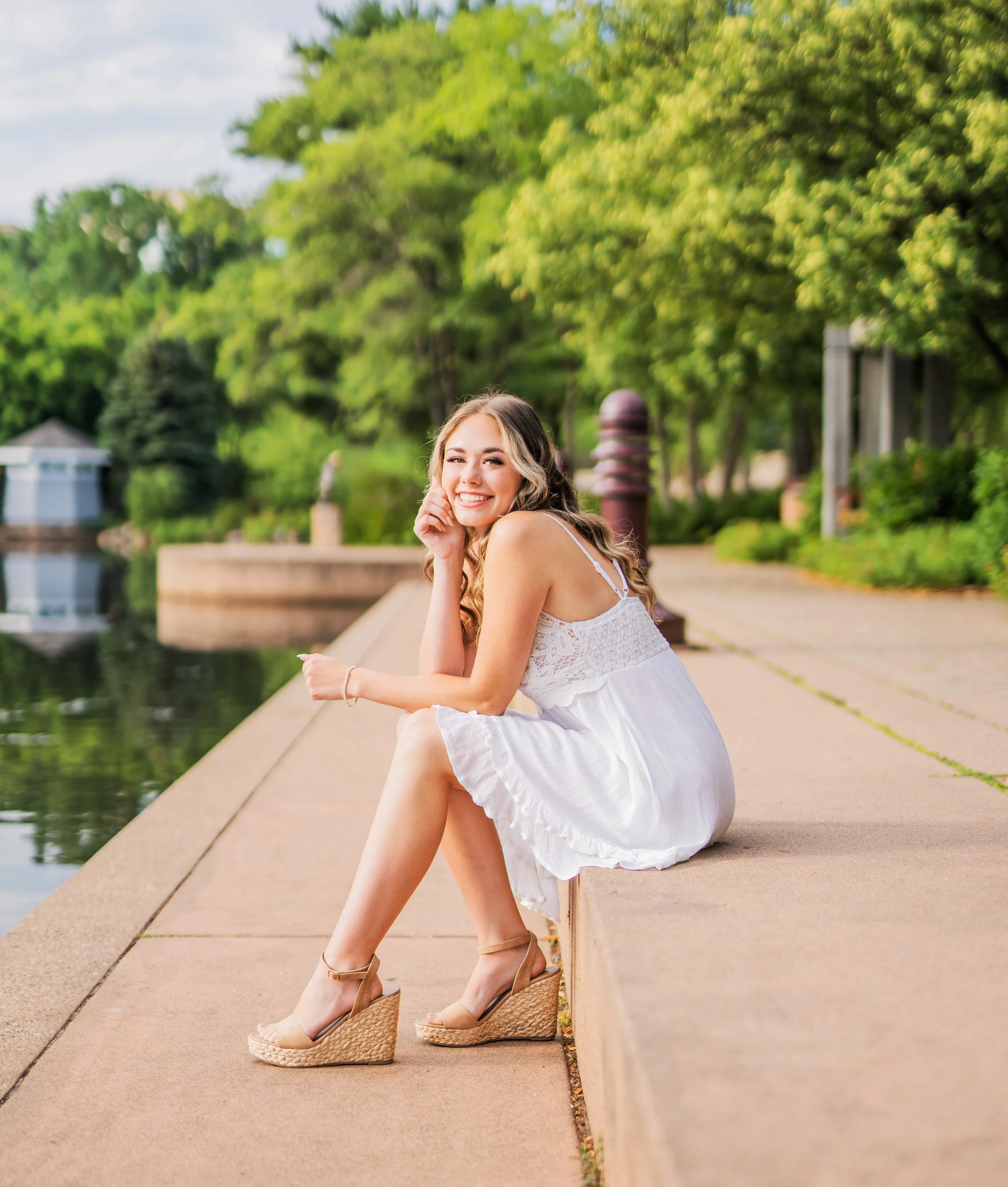 A young woman in a white dress and beige wedge sandals sitting on a concrete ledge by a body of water, smiling at the camera, with green trees and a park scene in the background.