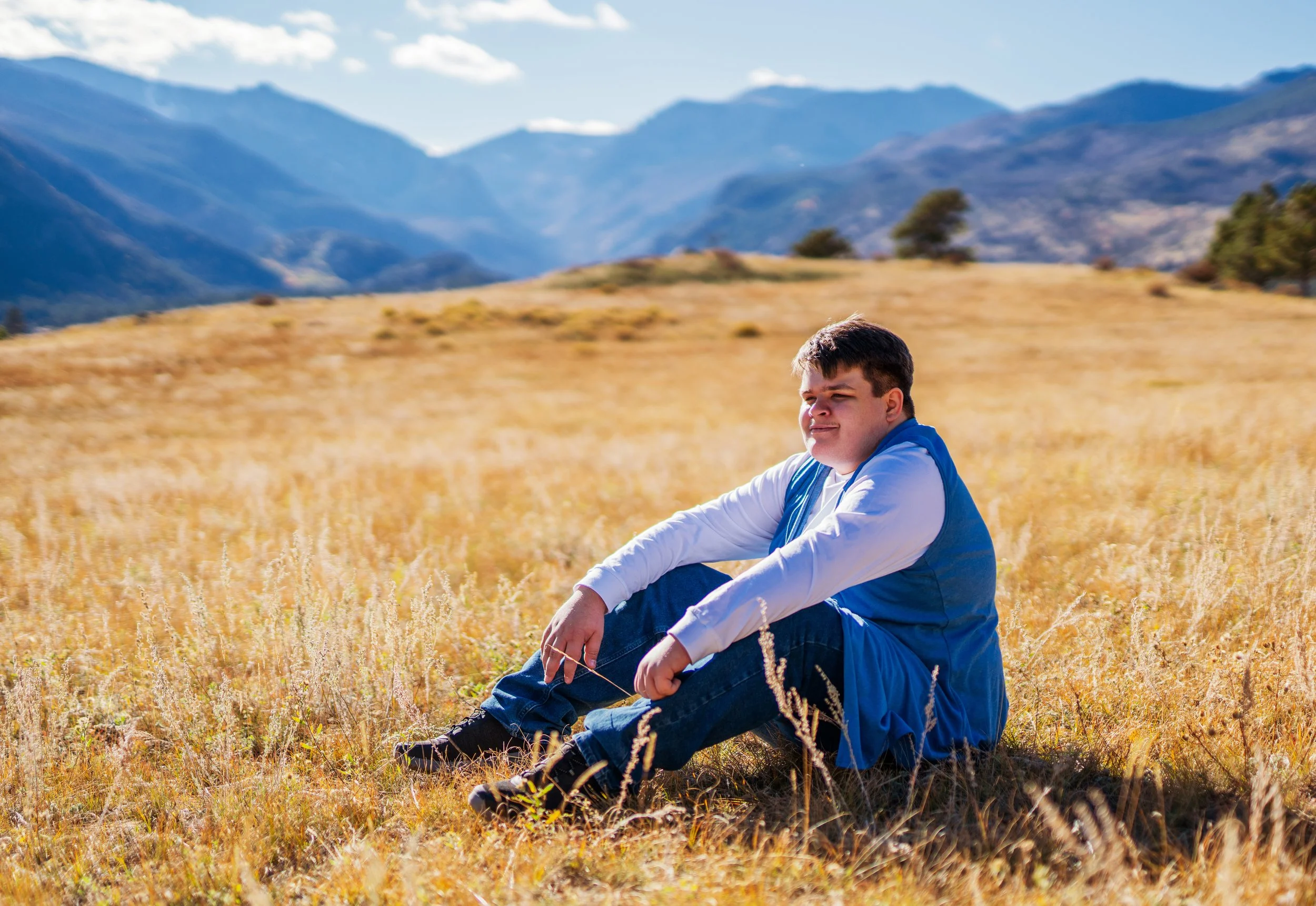 A young man sitting in a field of golden grass with mountain ranges and blue sky in the background.