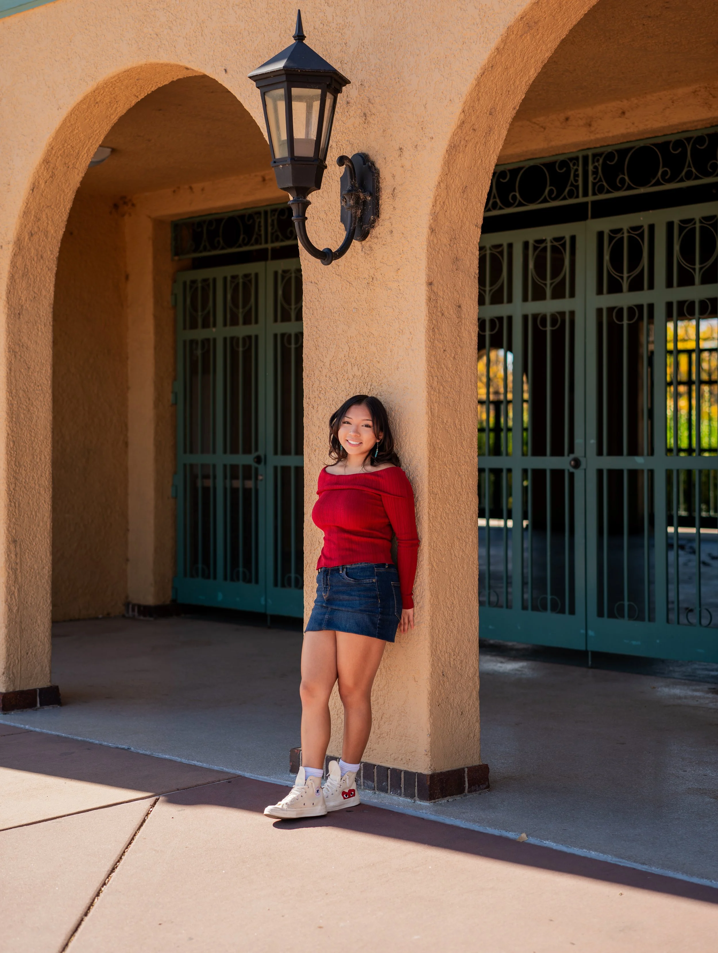 A young woman with dark hair in a red off-shoulder top, denim mini skirt, and white sneakers standing under an archway with teal gates and a black lantern on a beige wall.