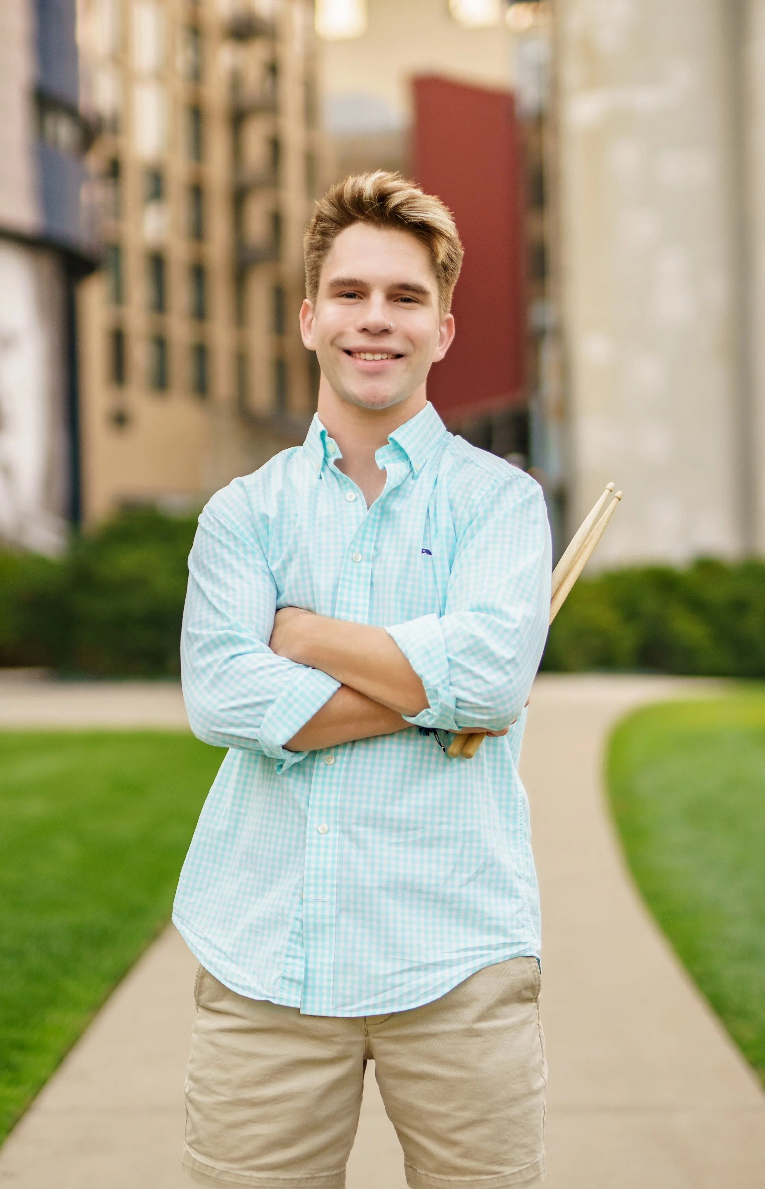 A young man with light skin, wearing a light blue checkered button-up shirt with beige shorts, smiling with arms crossed, standing outdoors on a pathway in a park or campus environment with grass and trees, holding a pair of drumsticks behind his bac