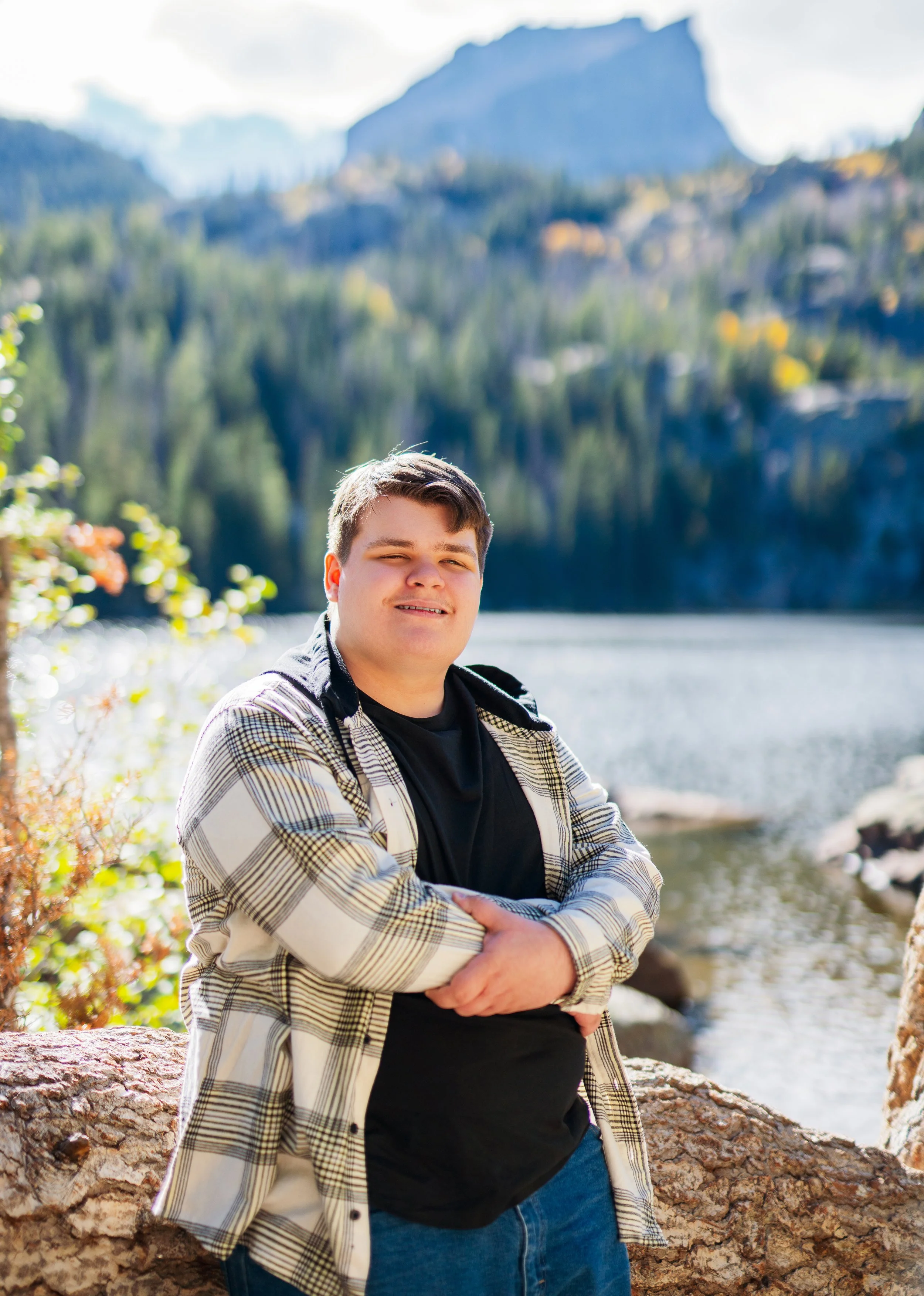A young man standing outdoors by a river with a mountain and forest in the background, smiling with his arms crossed, wearing a black t-shirt and plaid shirt.
