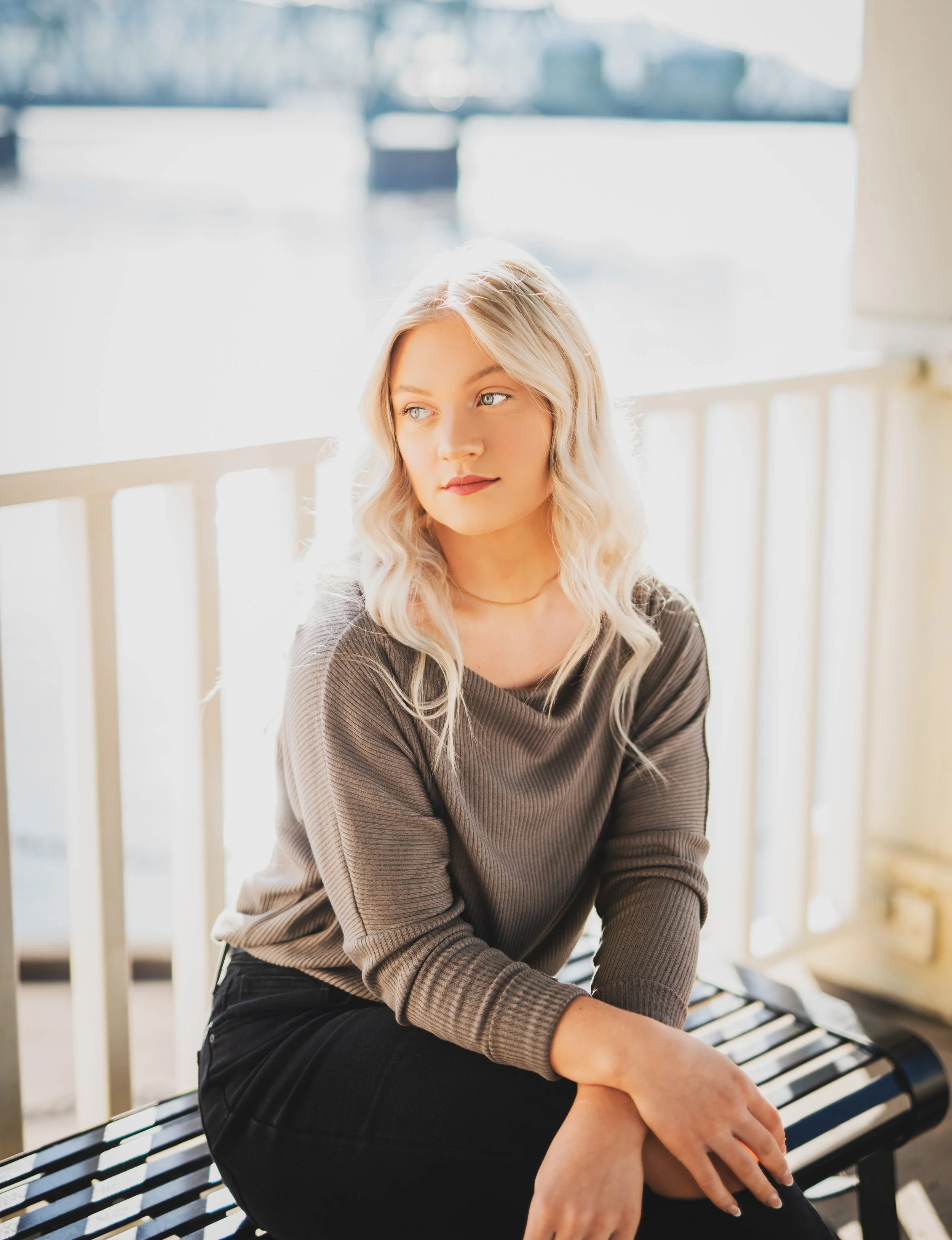 A young woman with blonde, wavy hair and blue eyes sitting on a bench near a window with a blurred water view in the background.
