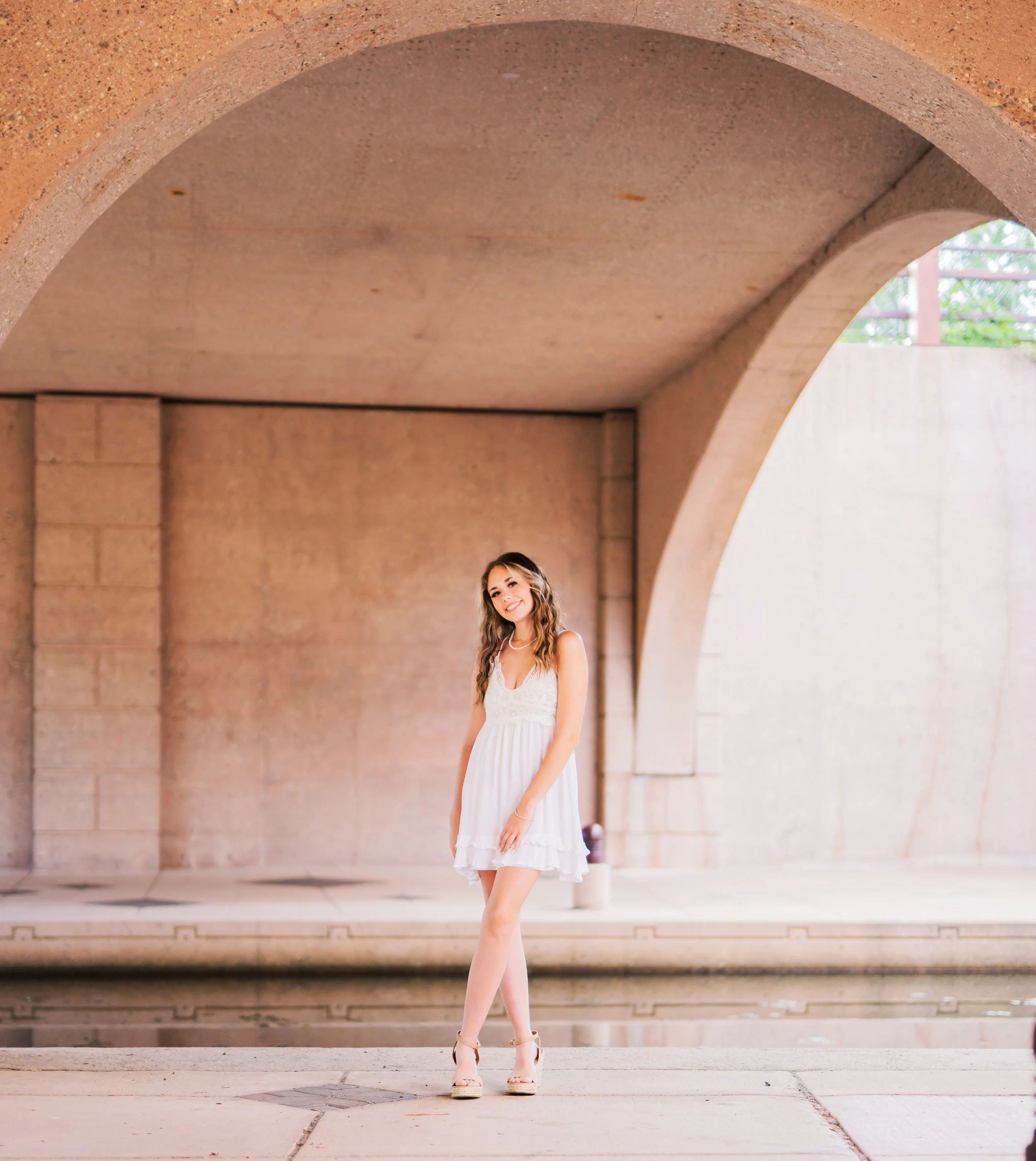 A young woman in a white dress standing under an archway with a fountain and concrete structure in the background.