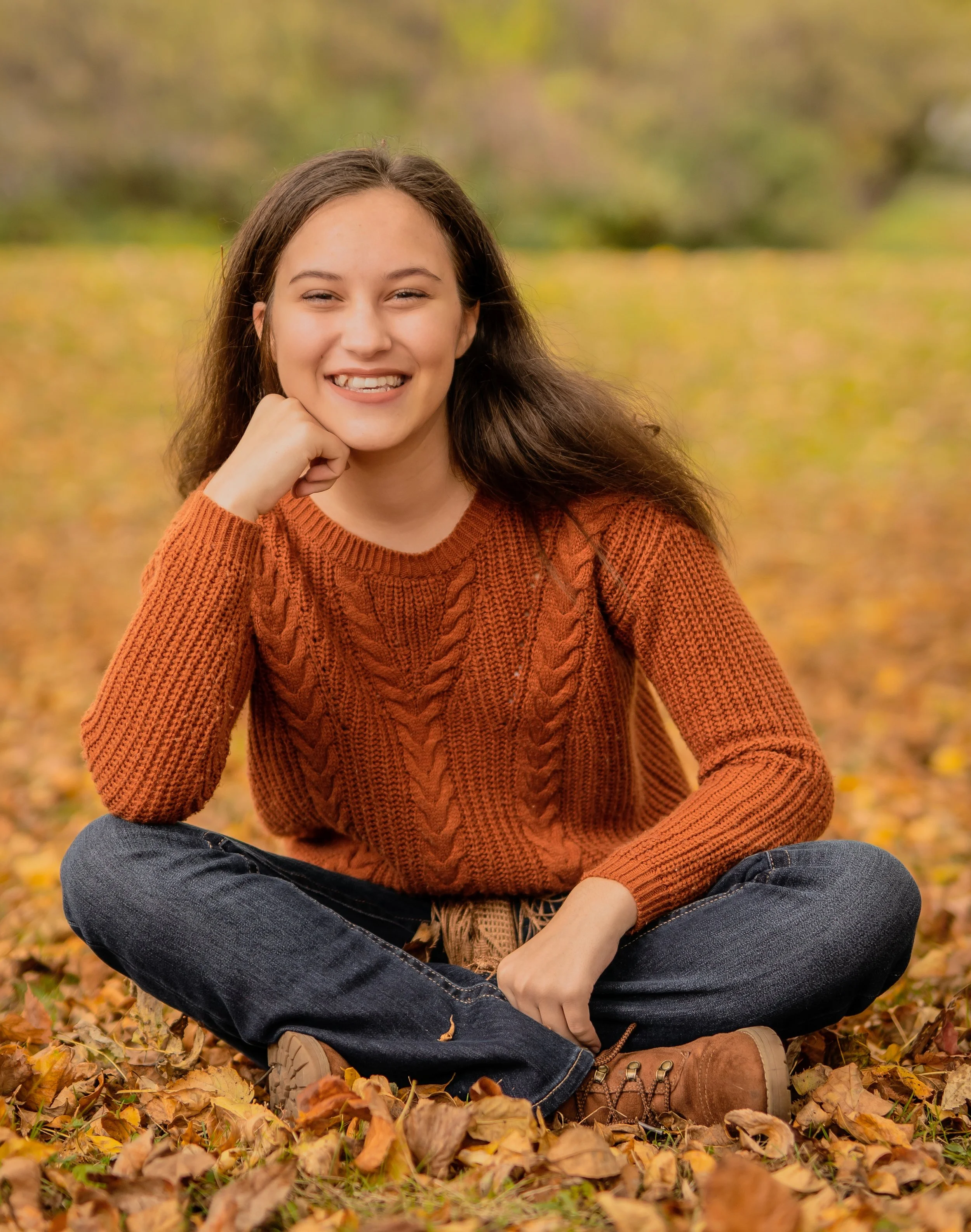 A young woman sitting cross-legged outdoors amidst fallen leaves, smiling, wearing a rust-colored sweater, jeans, and brown boots, with an autumnal background.