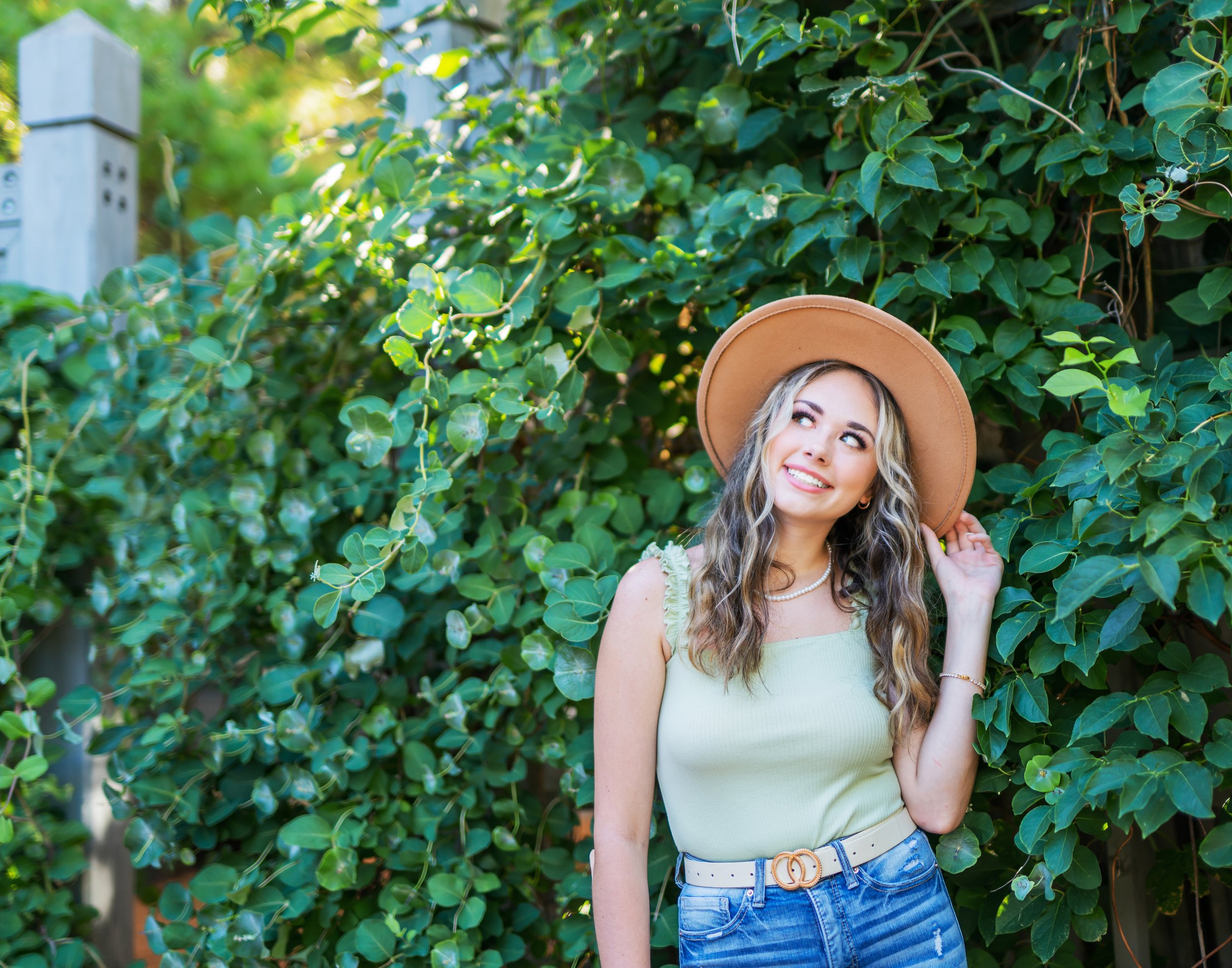 A young woman with long wavy hair in a green tank top and jeans, wearing a wide-brimmed hat, standing in front of green leafy bushes, smiling and touching her hat with one hand.