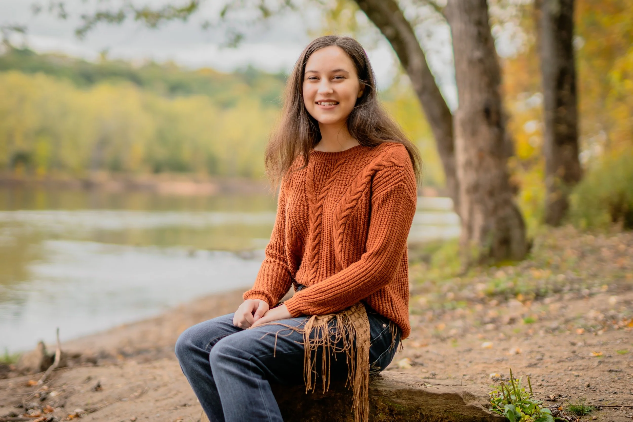 A young woman with long brown hair, wearing an orange sweater and jeans, sitting on a log near a river in a park during fall, with trees and colorful leaves in the background.