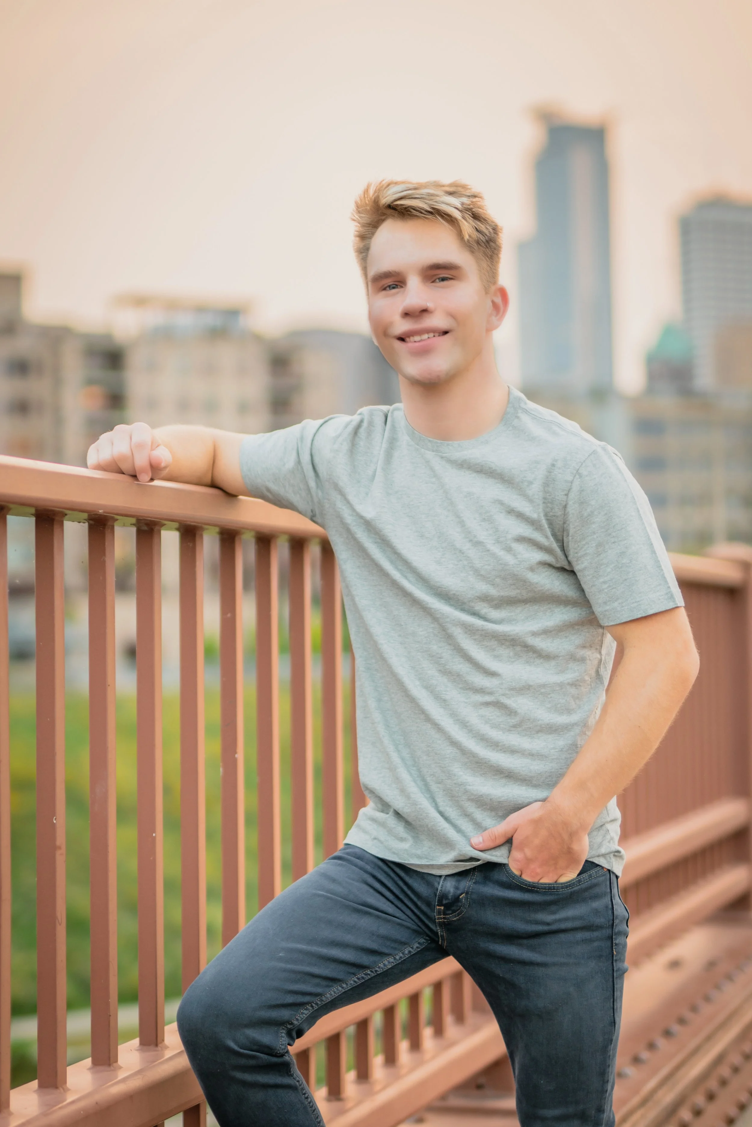 A young man leaning on a railing on a bridge, smiling at the camera, with city buildings in the background during sunset.