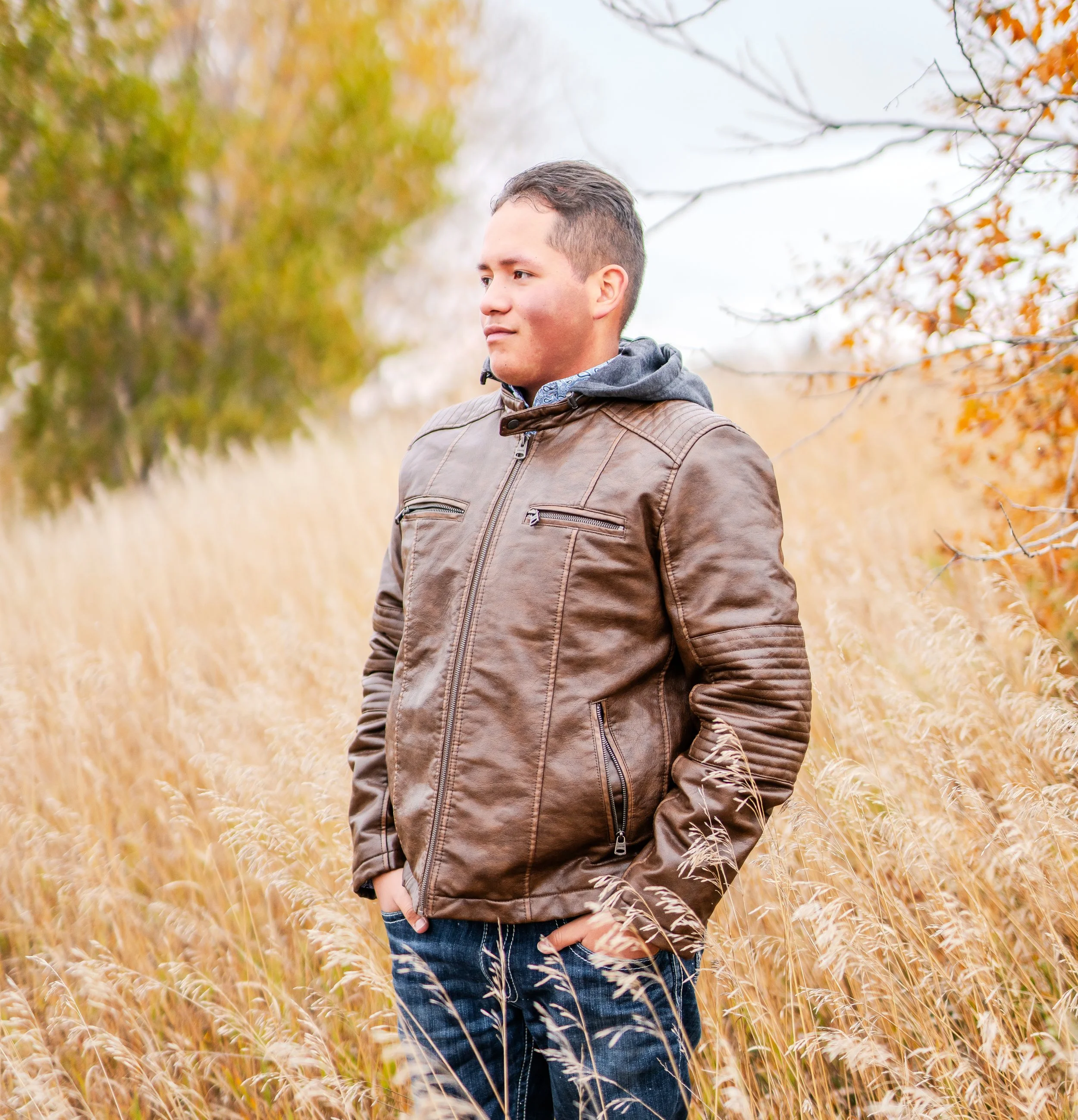 A young man in a brown leather jacket stands in a field with tall, golden grasses, autumn trees, and overcast sky, looking to the side.