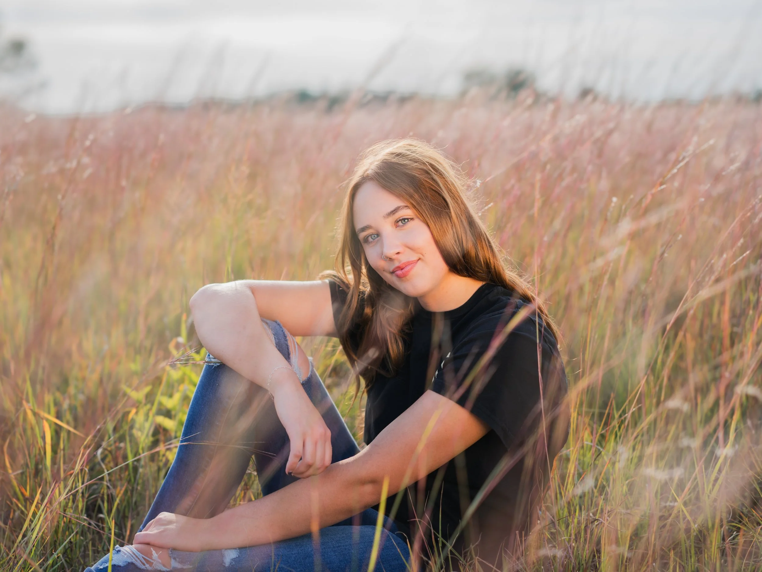 Young woman sitting in a field of tall grass, smiling at the camera during sunset.