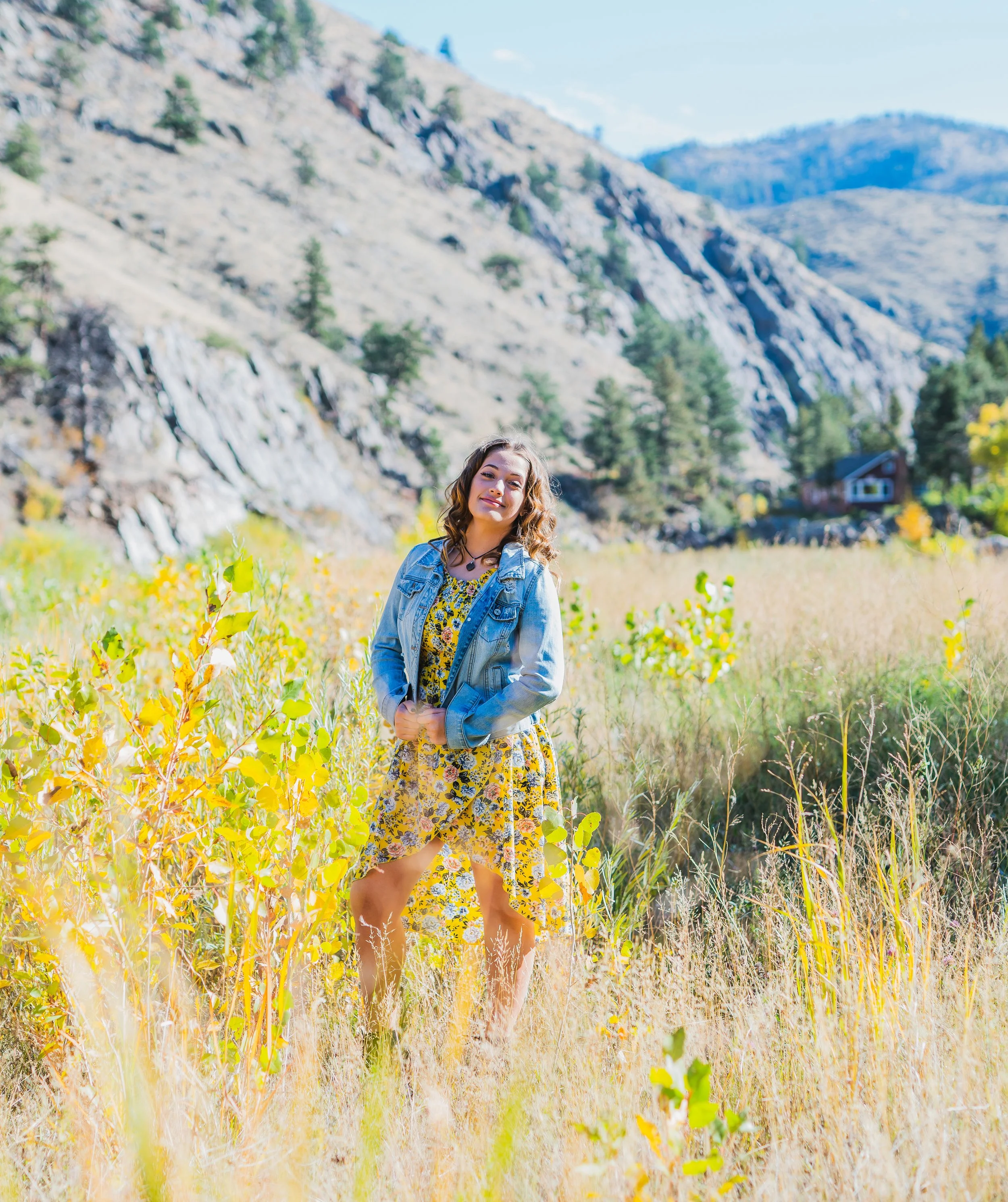 A woman standing in a field of tall grasses and yellow flowers with mountains and forested hills in the background on a sunny day.