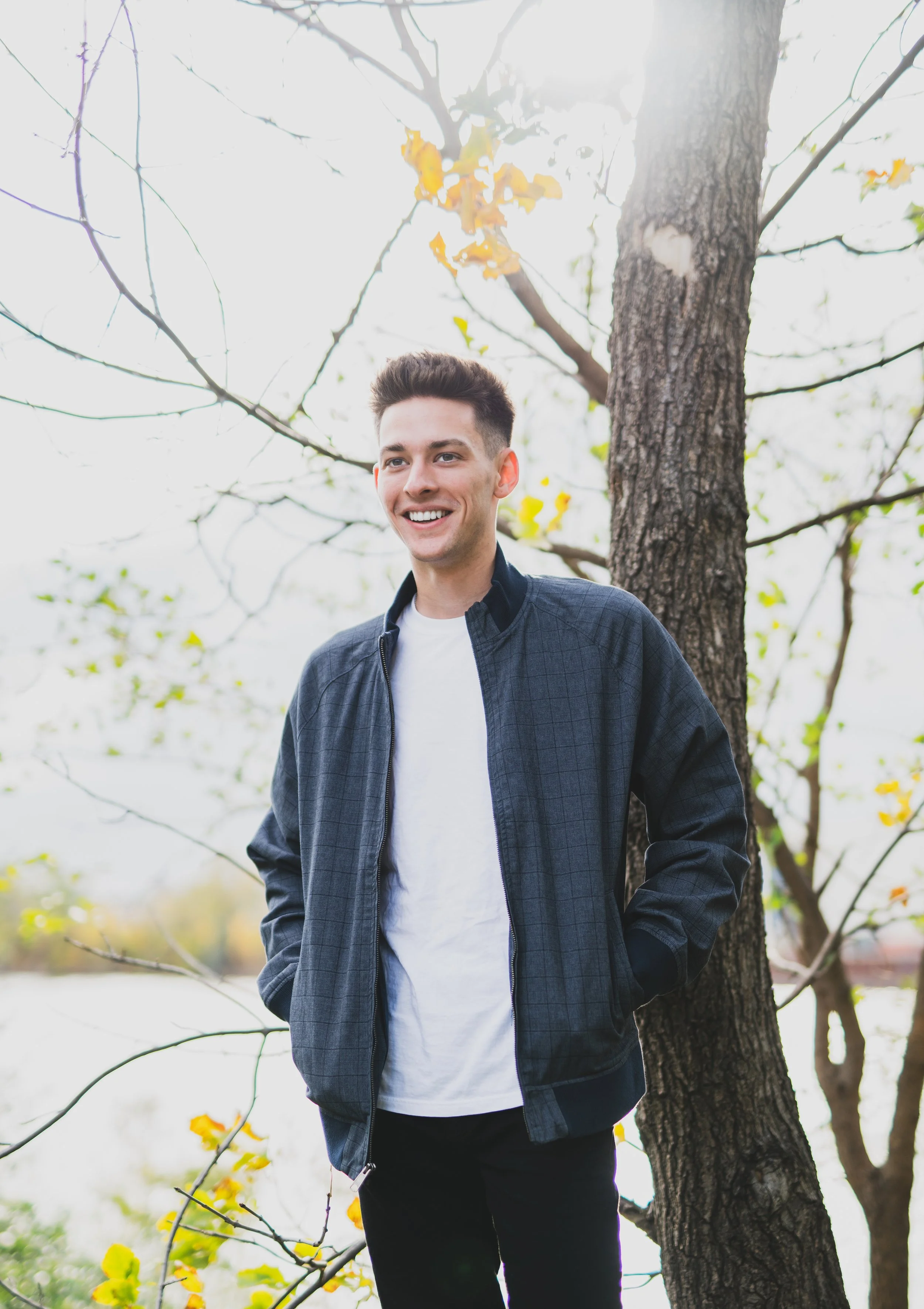 A young man with short dark hair smiling and standing outdoors near a tree during autumn.