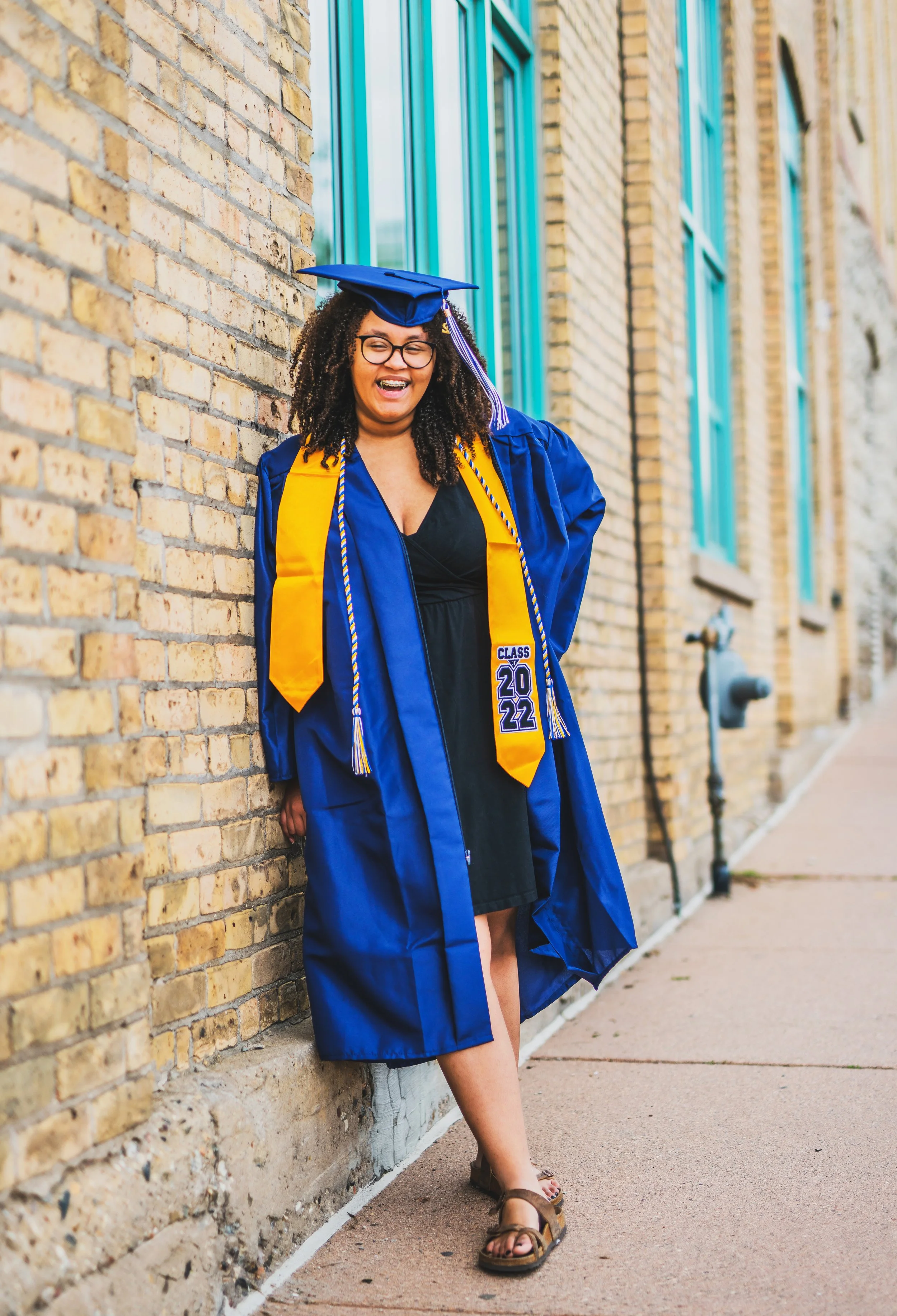 A woman in a graduation cap and gown poses outdoors against a brick wall, smiling and looking joyful.