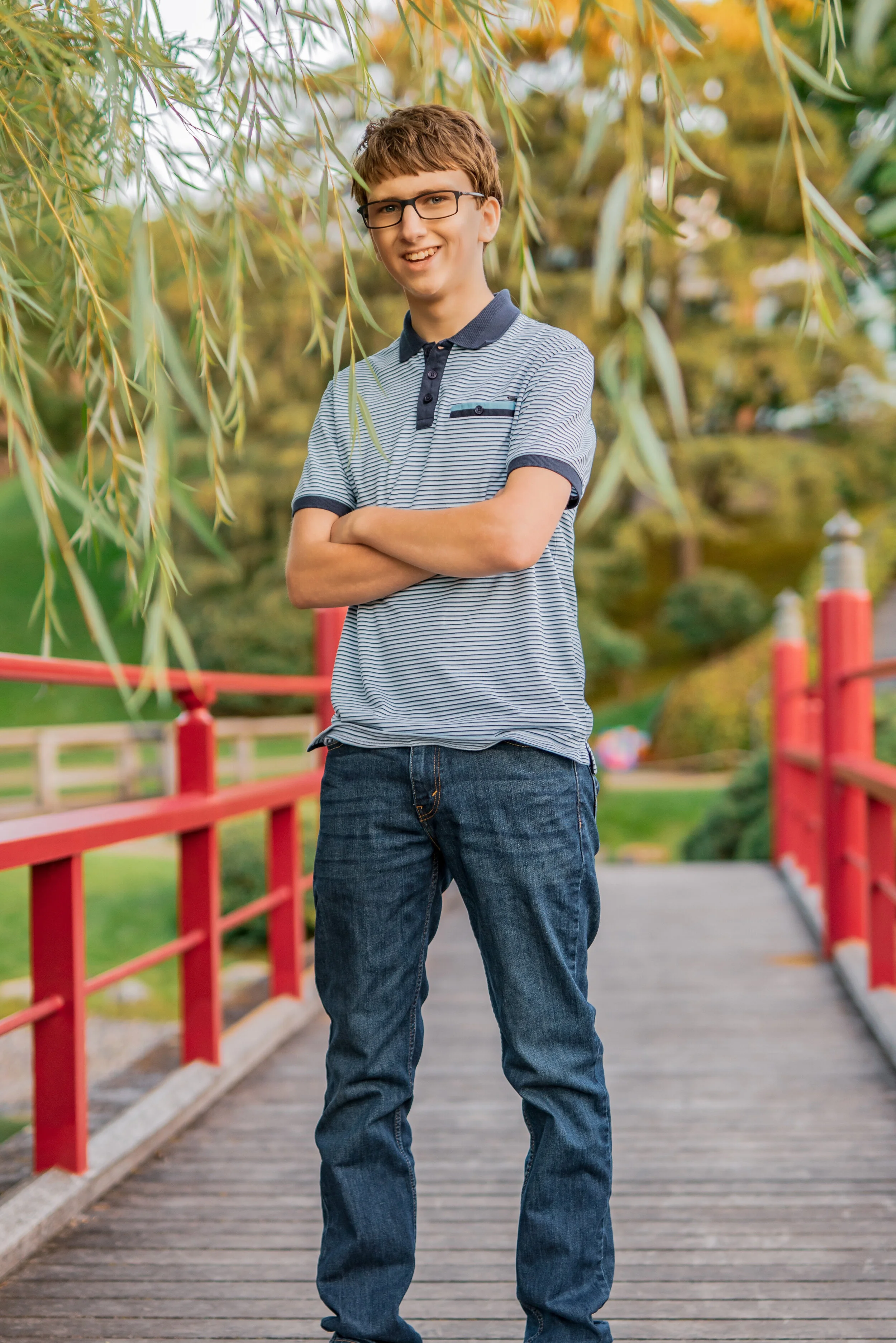 A teenage boy wearing glasses, a striped polo shirt, and jeans standing on a wooden bridge with a red railing in a park, smiling with arms crossed in front of a background of green trees and plants.