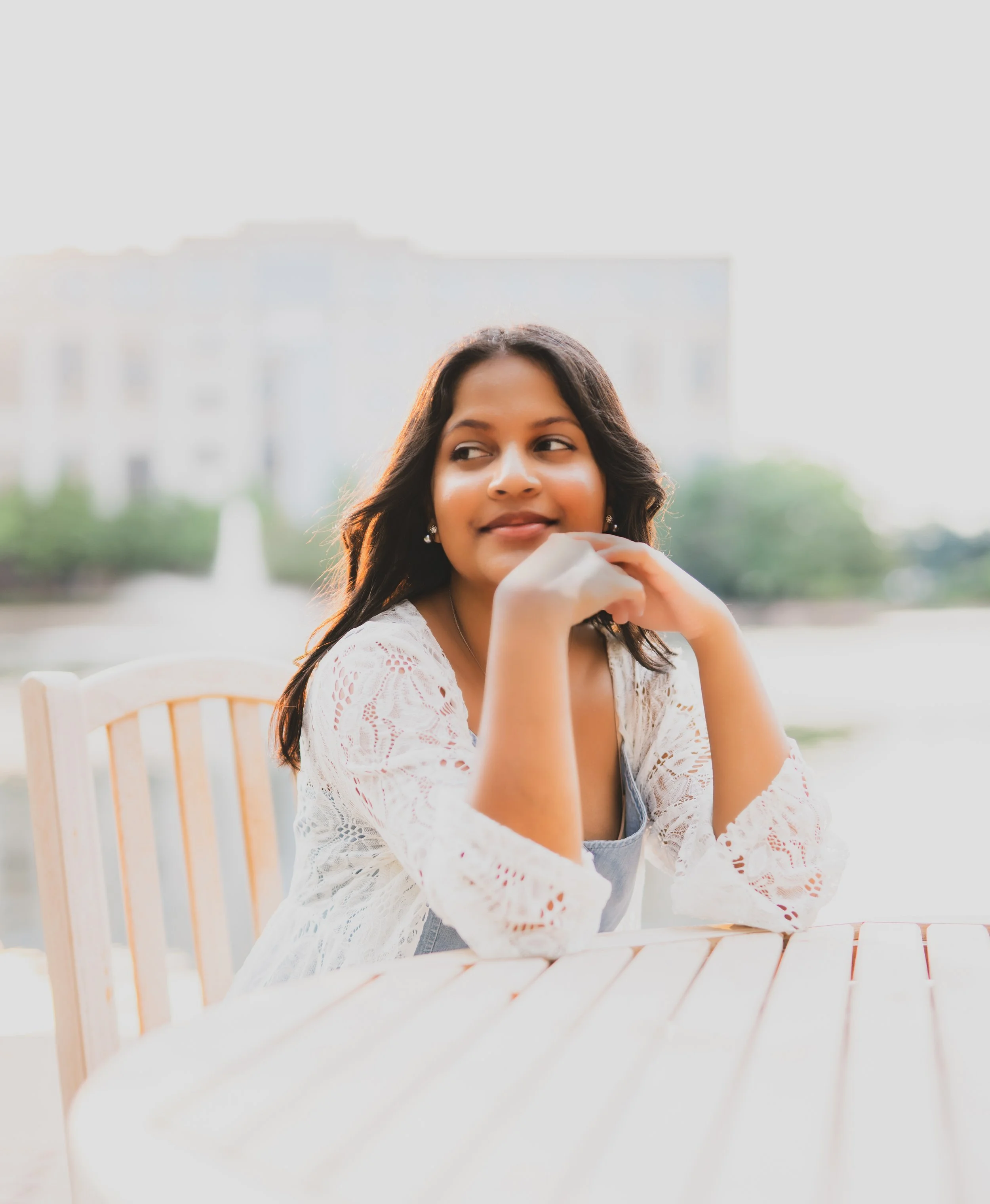 A young woman sitting at a table outdoors with a fountain and a building in the background, smiling softly and looking to the side.