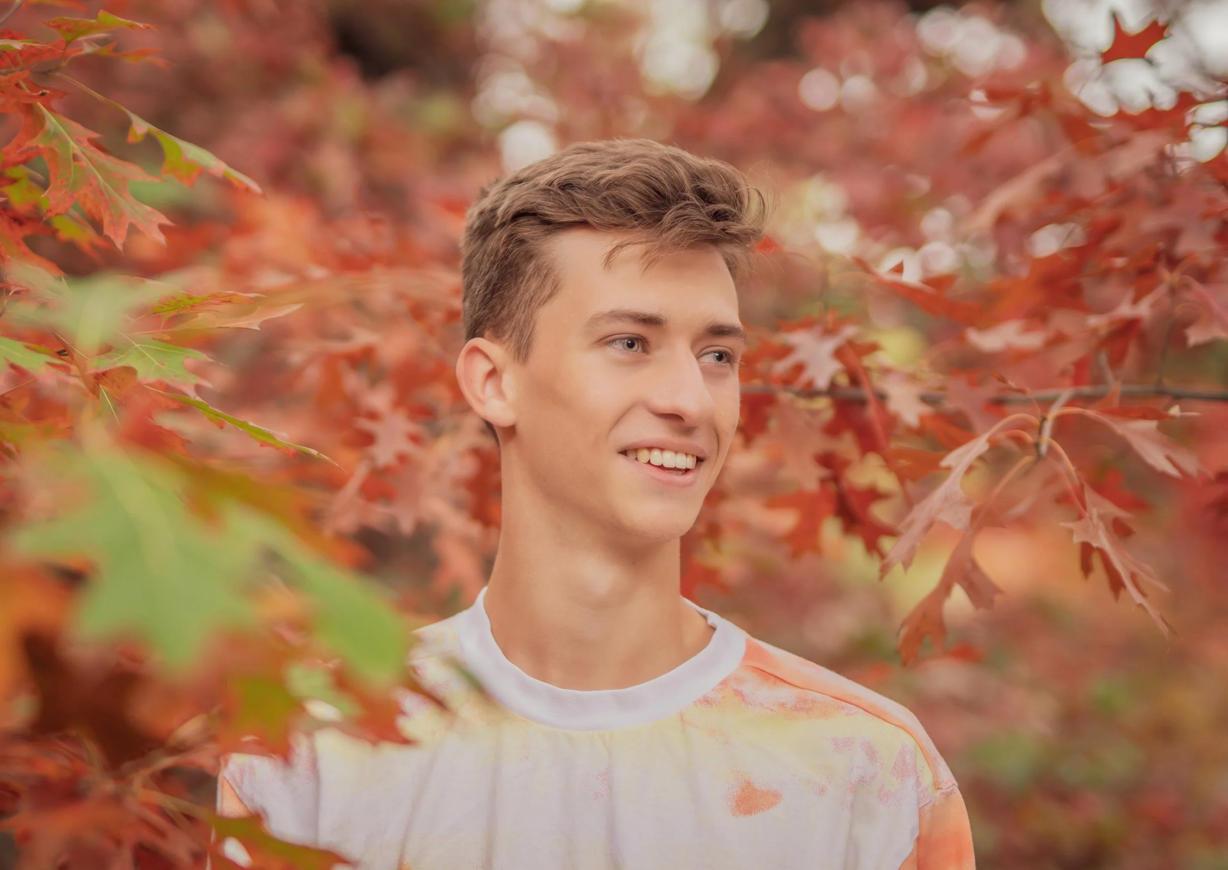 A young man smiling outdoors among autumn red and green leaves.