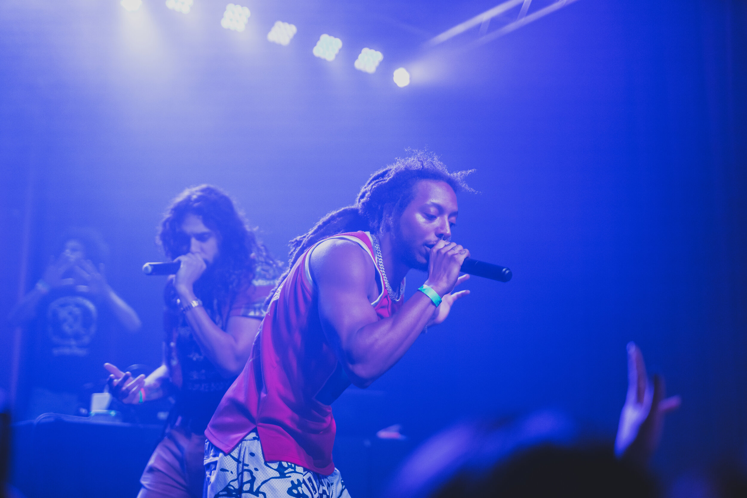 Performers on stage with microphones under blue and purple stage lights, with audience hand visible in foreground.
