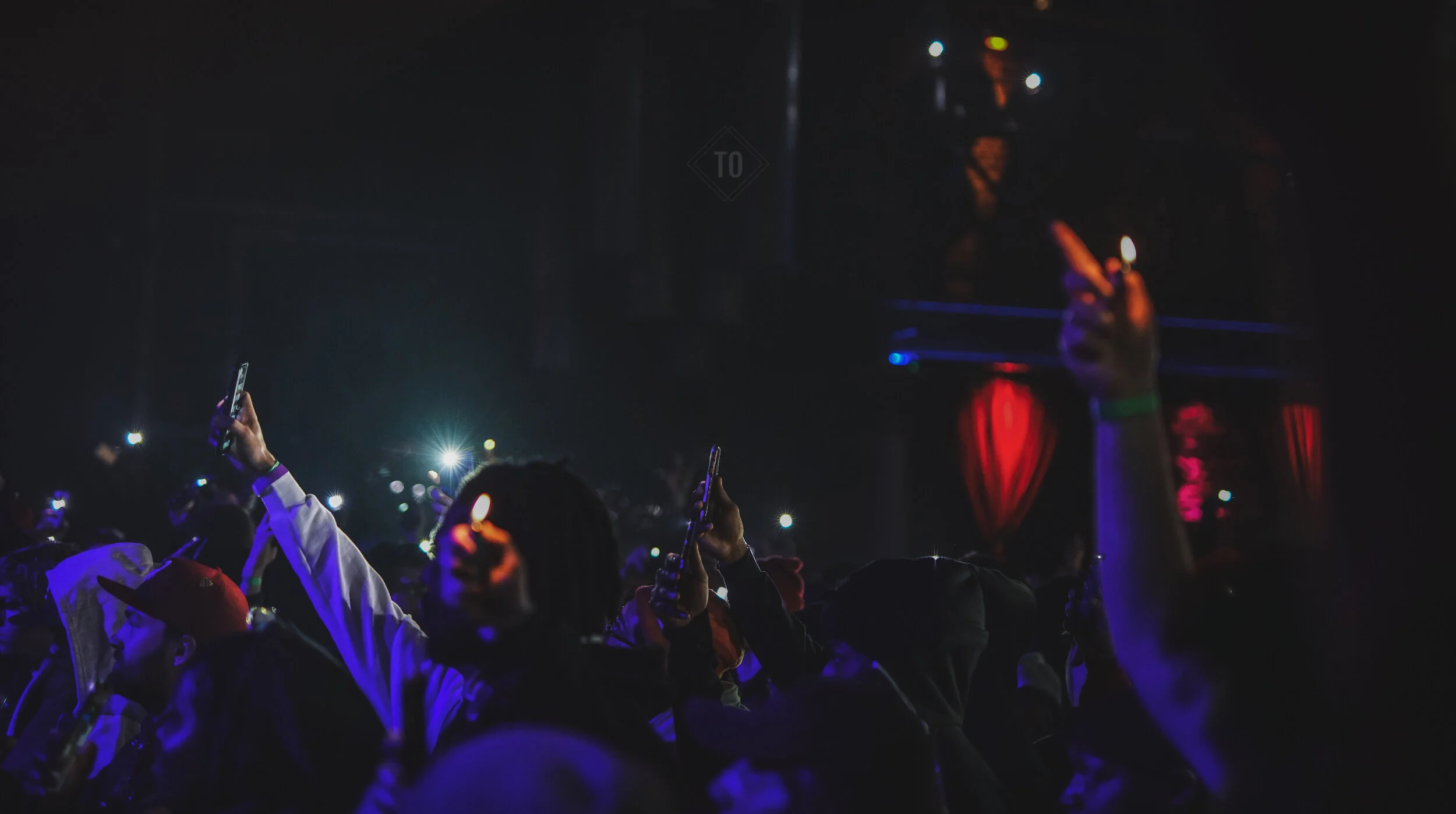 Crowd at a nighttime concert or event, illuminated by their smartphone screens, with some hands raised, and a stage with red curtains in the background.