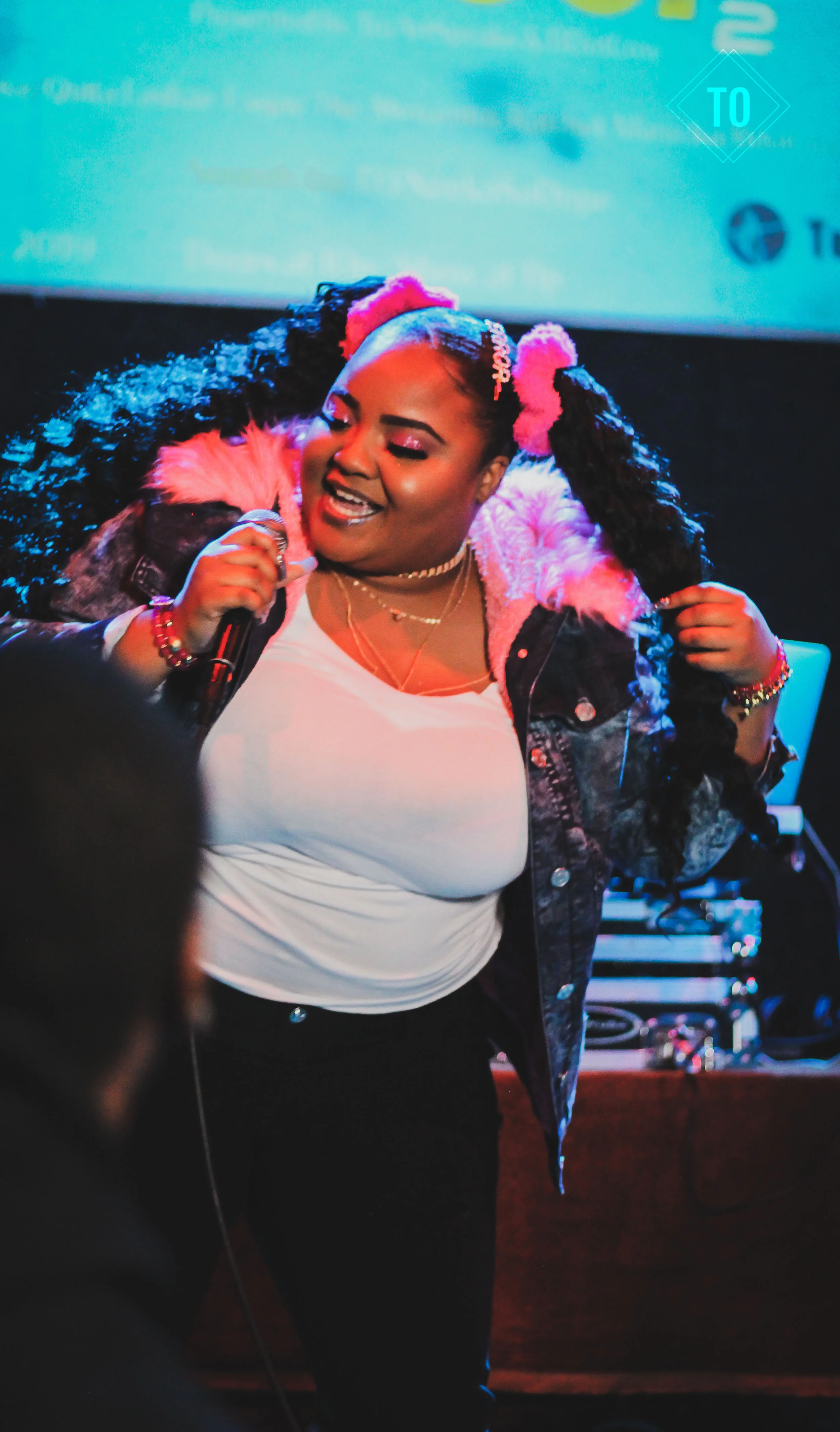 A woman with curly hair tied with pink scrunchies, wearing a denim jacket with pink fur lining, singing or speaking into a microphone at an indoor event.