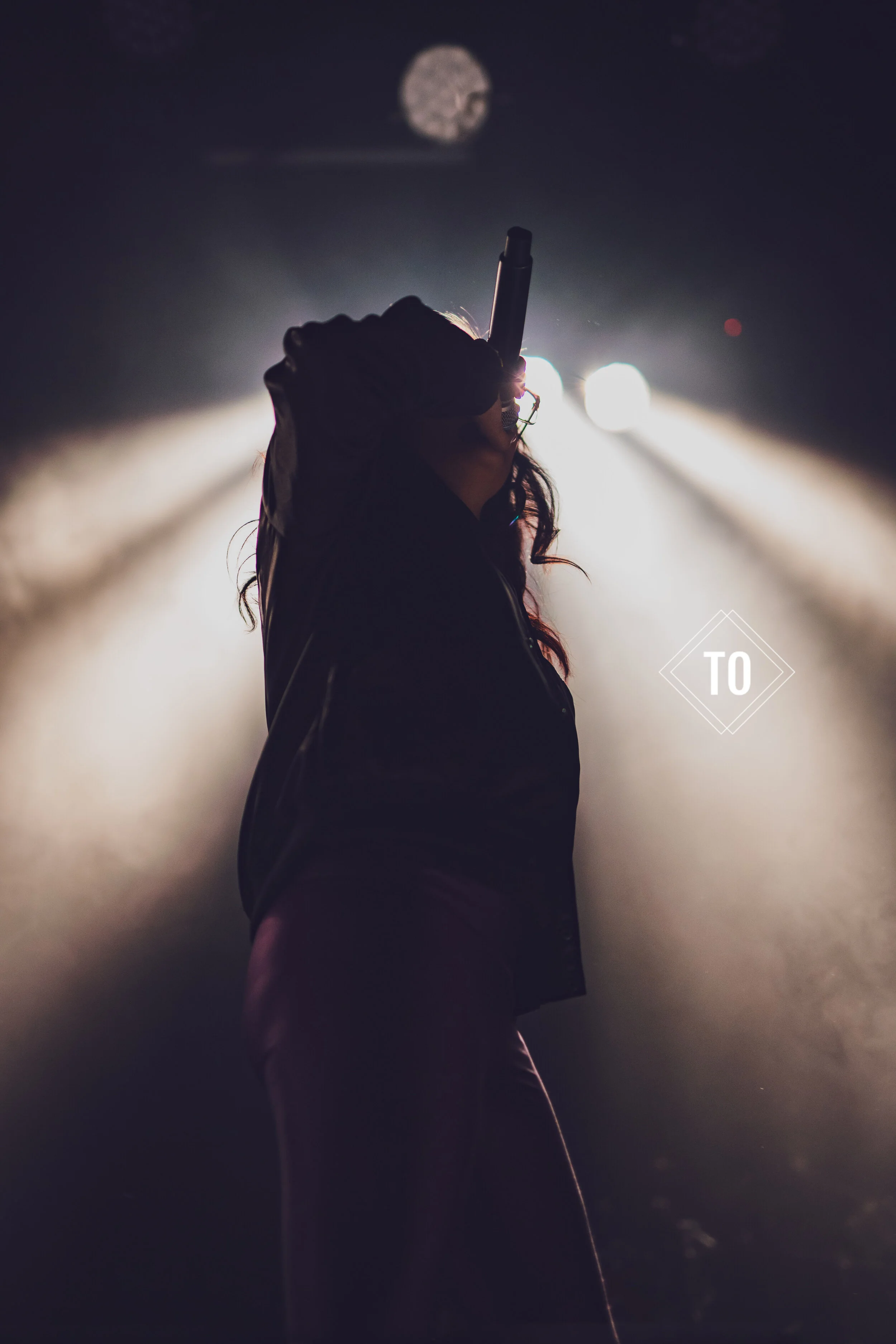 A woman performing on stage at night, holding a microphone and facing away from the camera, with bright stage lights shining behind her and a full moon in the sky.