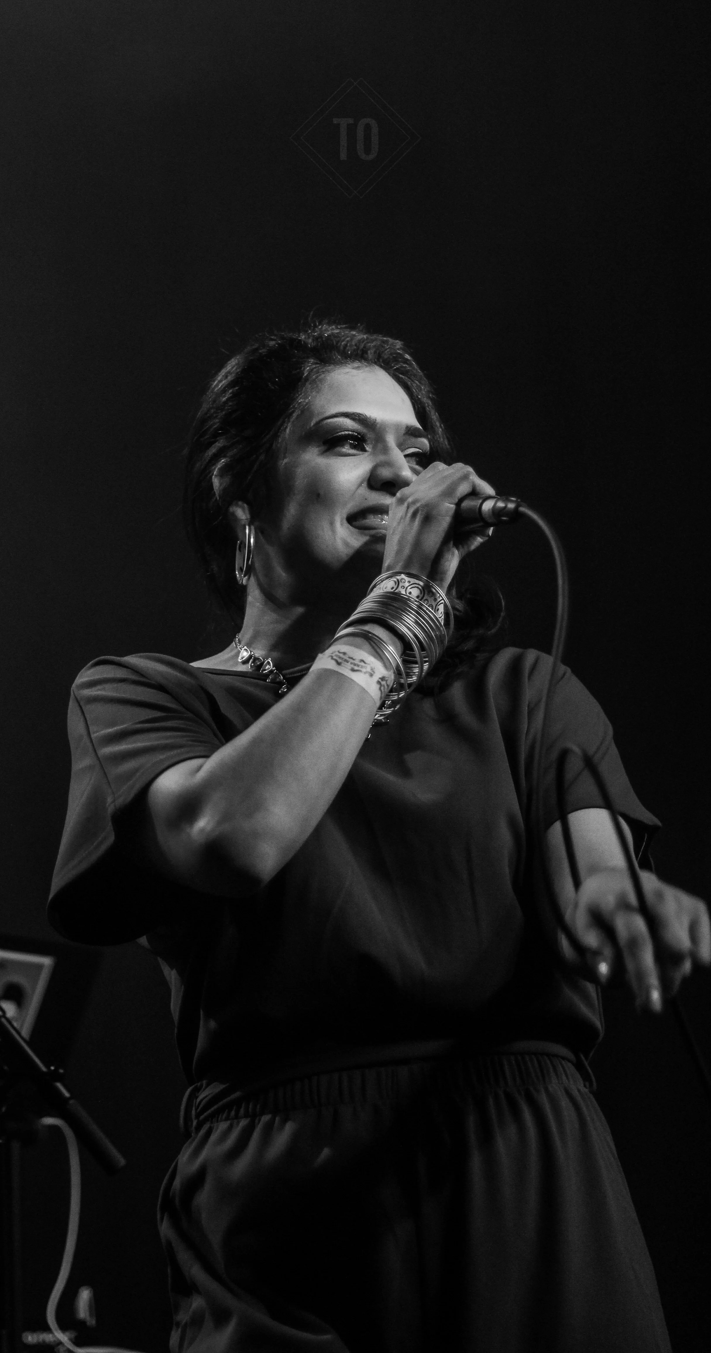 Black and white photo of a woman singing into a microphone on stage, wearing a dark shirt, with bracelets and earrings, with a focused expression.