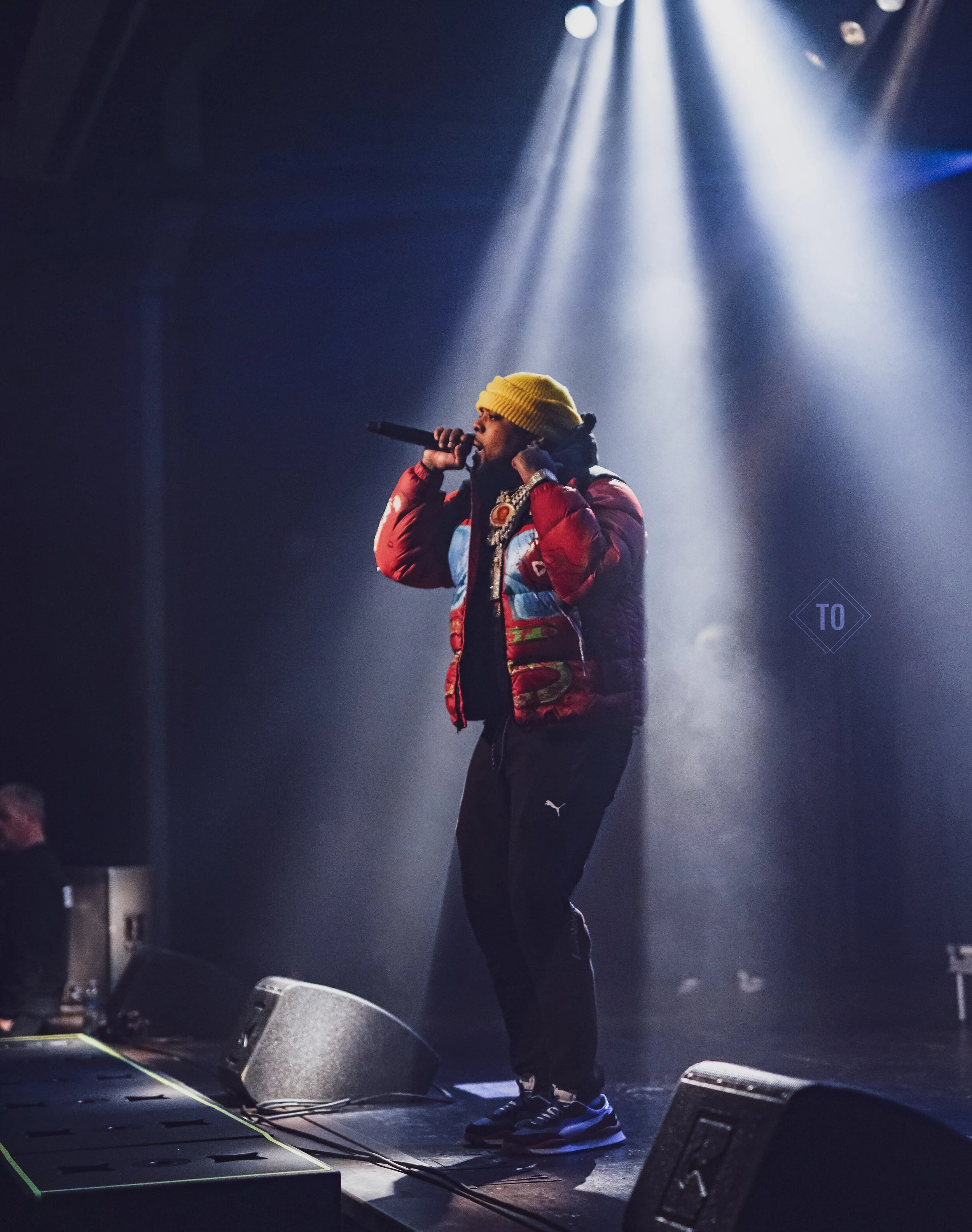 A person performing on a stage, holding a microphone and wearing a yellow beanie, a colorful jacket, black pants, and sneakers, illuminated by stage lights.