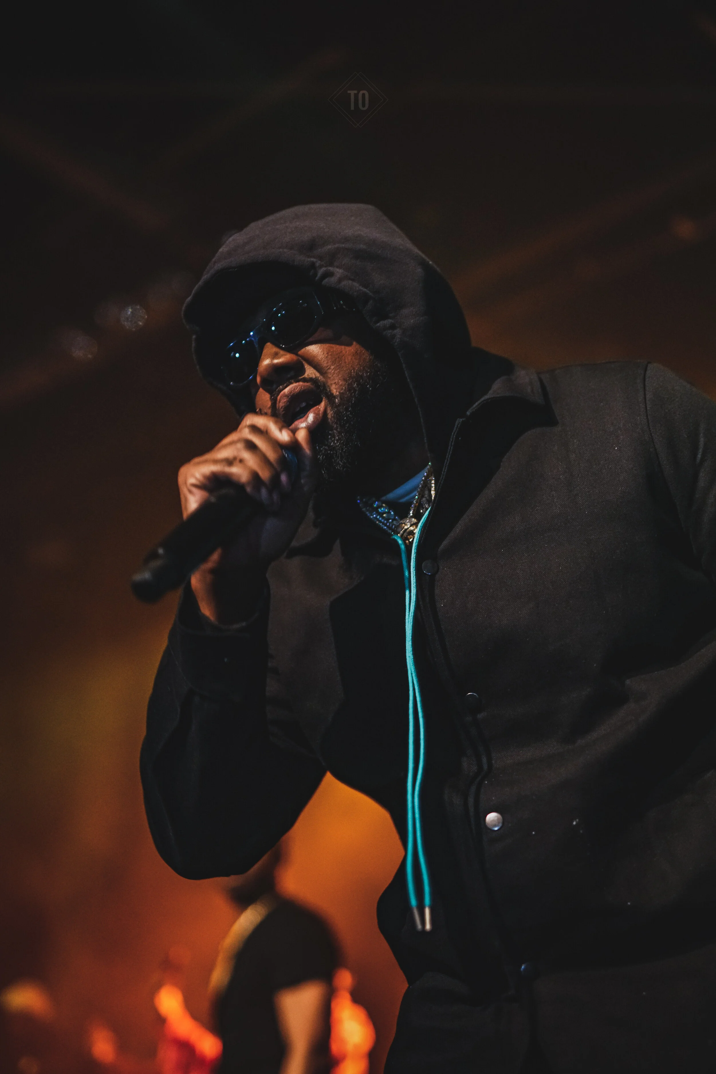 A man wearing dark sunglasses, a hoodie, and a black jacket, holding a microphone and performing on stage with warm lighting.