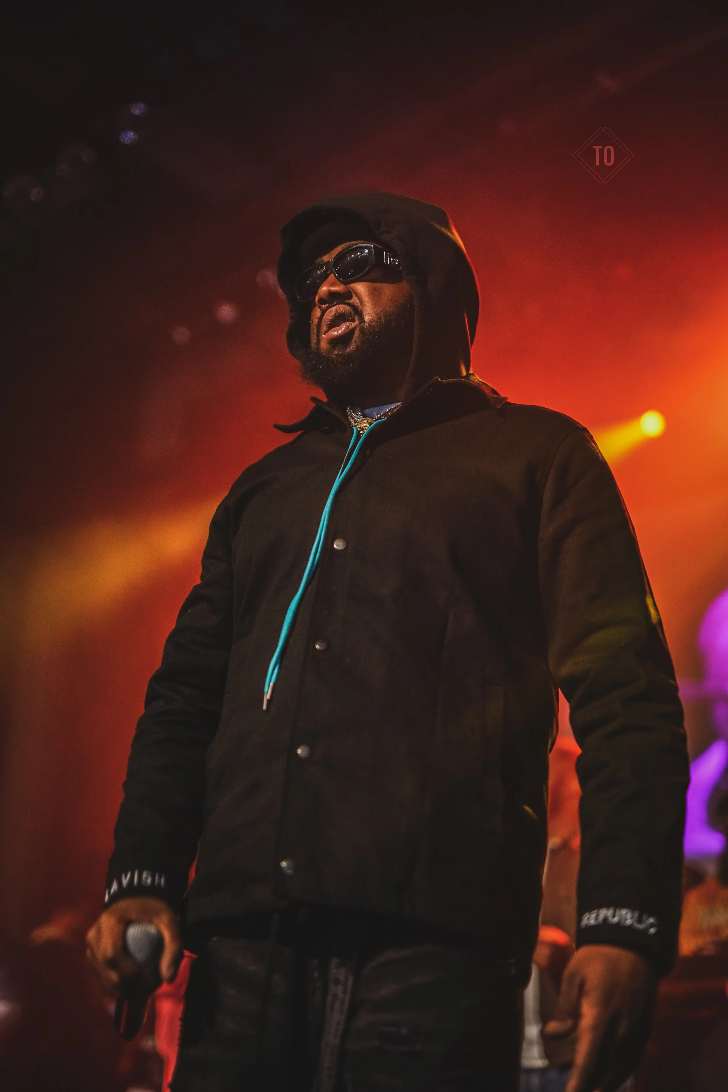 A man wearing a black jacket, sunglasses, and a hood, standing on a stage with colorful lights behind him.
