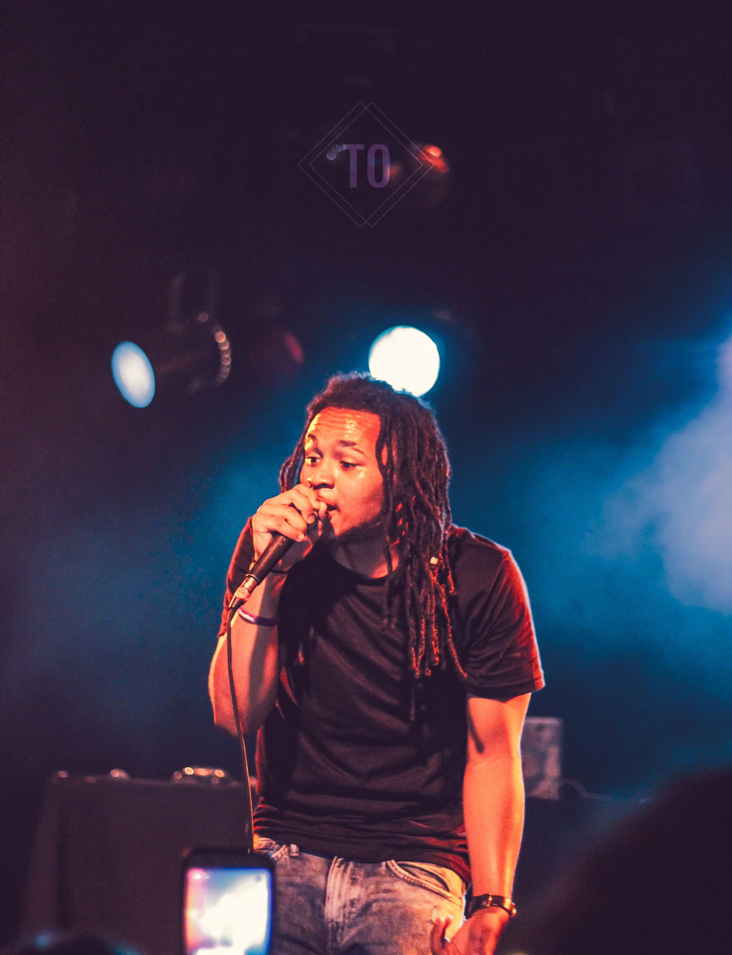 A male performer with dreadlocks singing into a microphone on stage under colorful stage lights.