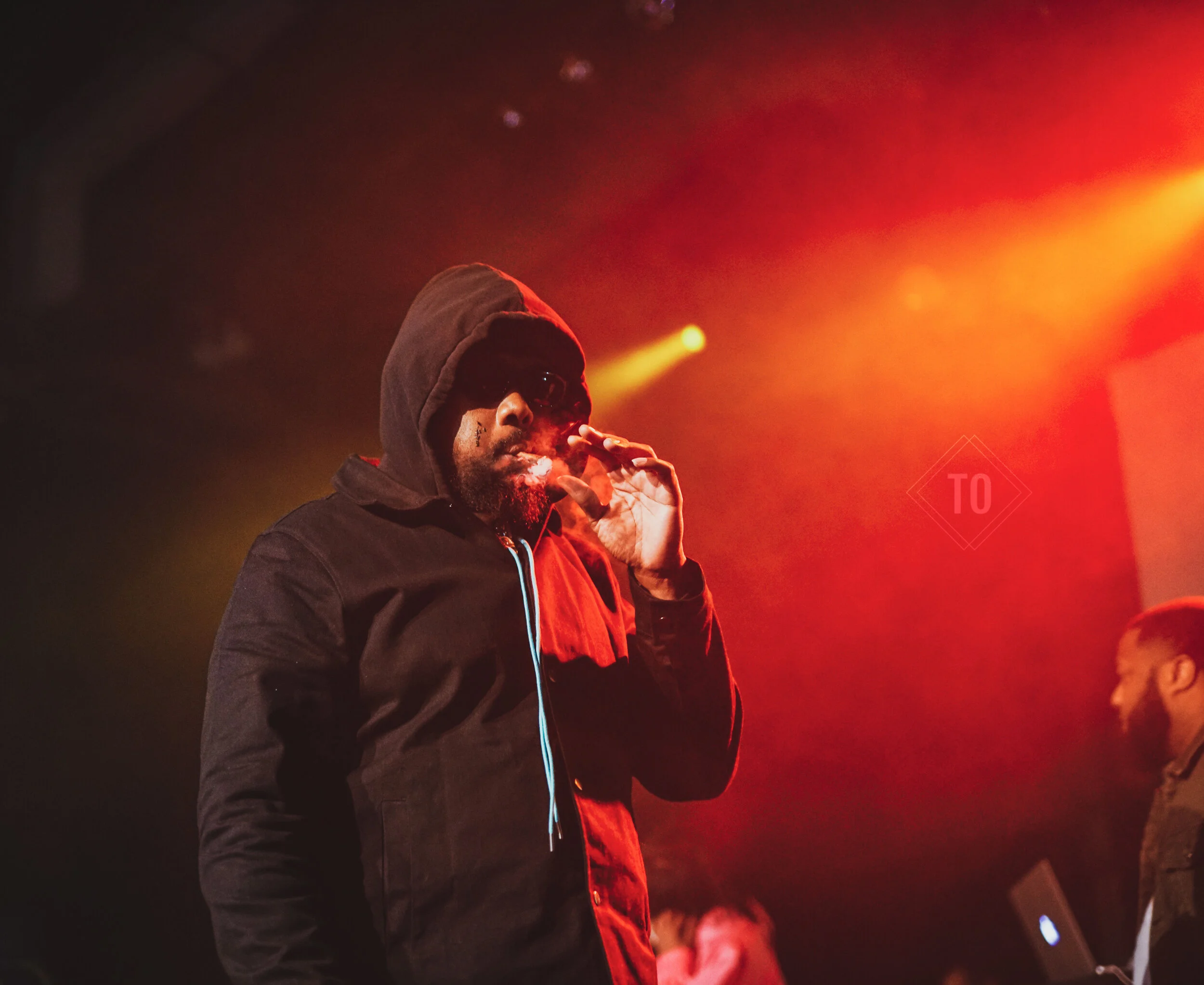 A man wearing a black hoodie, sunglasses, and smoking, standing in a dimly lit room with red and yellow stage lights.