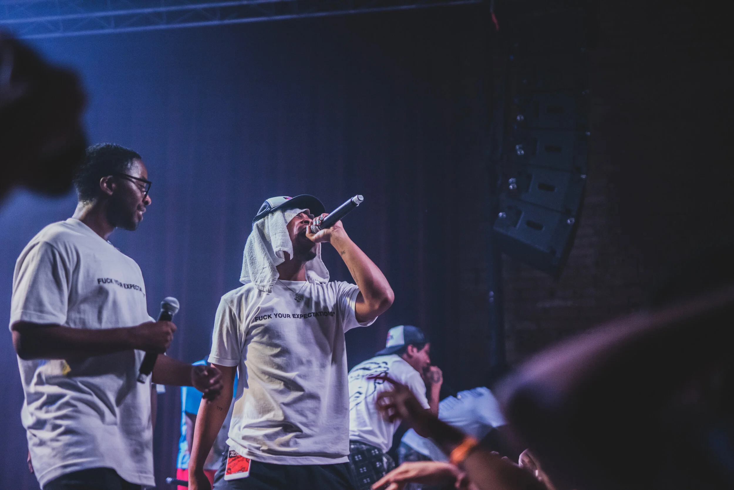 Group of performers on stage, with one singer wearing a cap and towel over his head, holding a microphone and singing, others holding microphones, in a dimly lit concert setting.