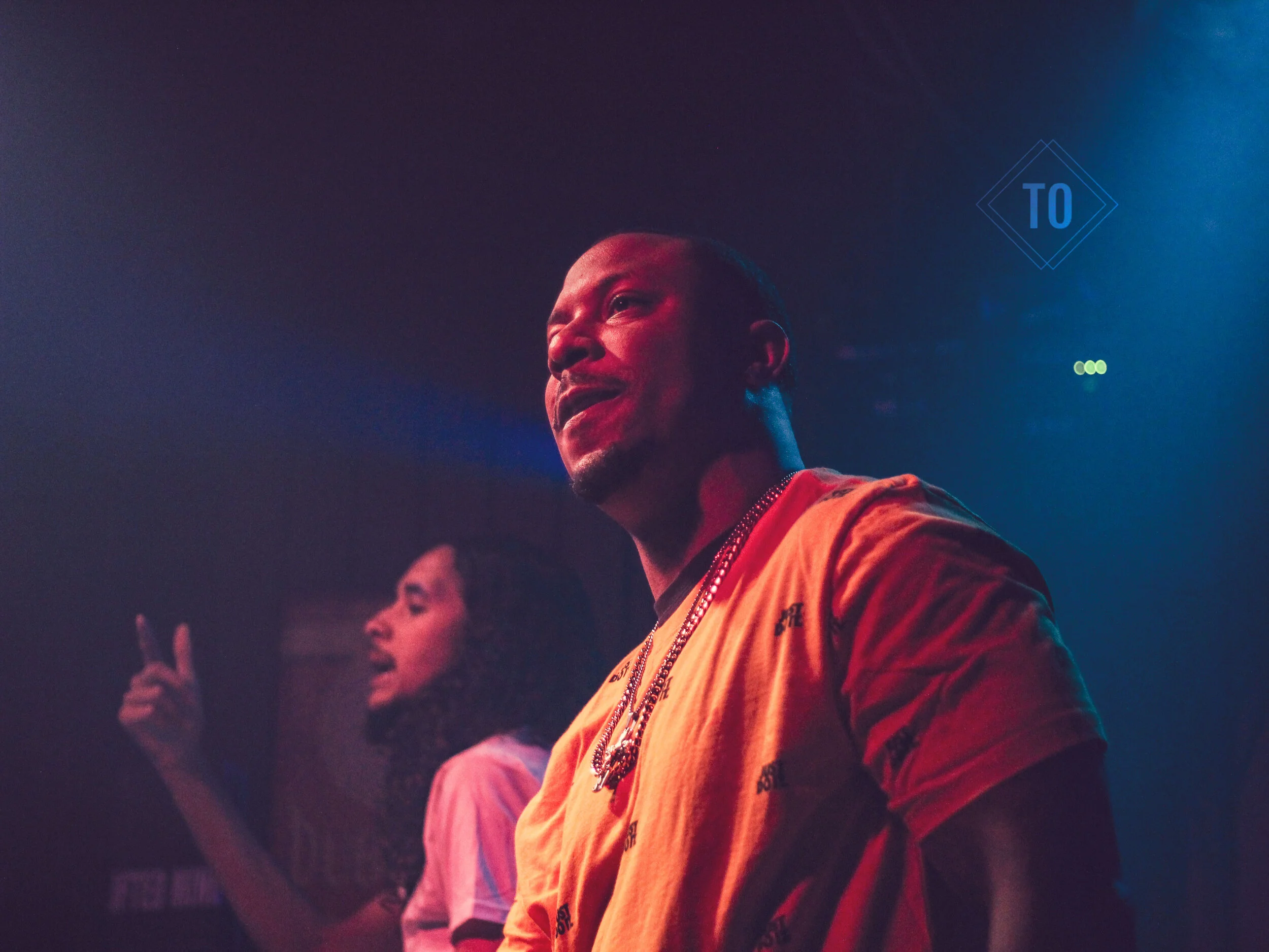 A man wearing a yellow shirt and chains is looking to the side under colorful stage lighting, with a woman in the background pointing upward.