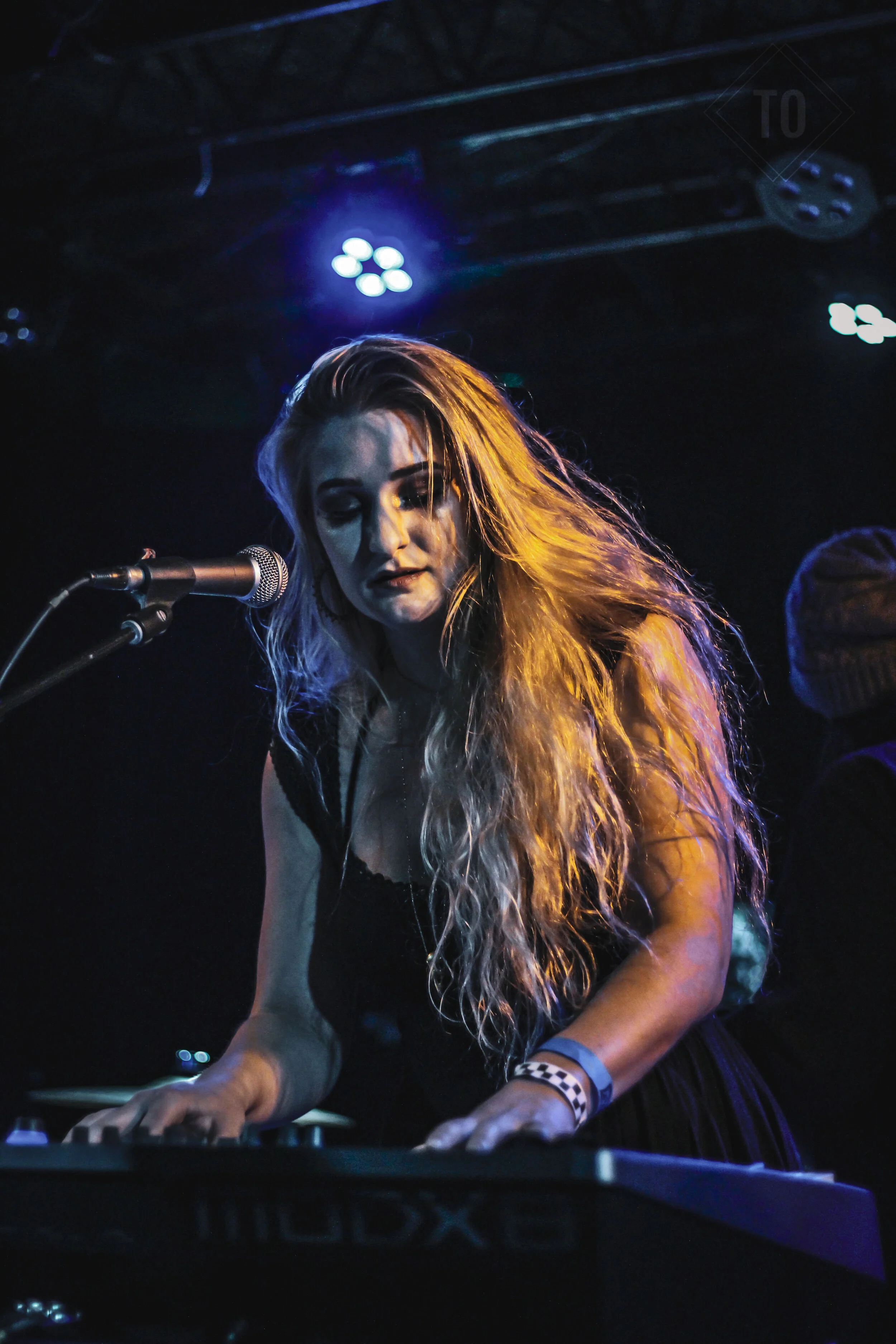 A female musician with long curly blonde hair playing a keyboard on stage, with stage lights overhead.