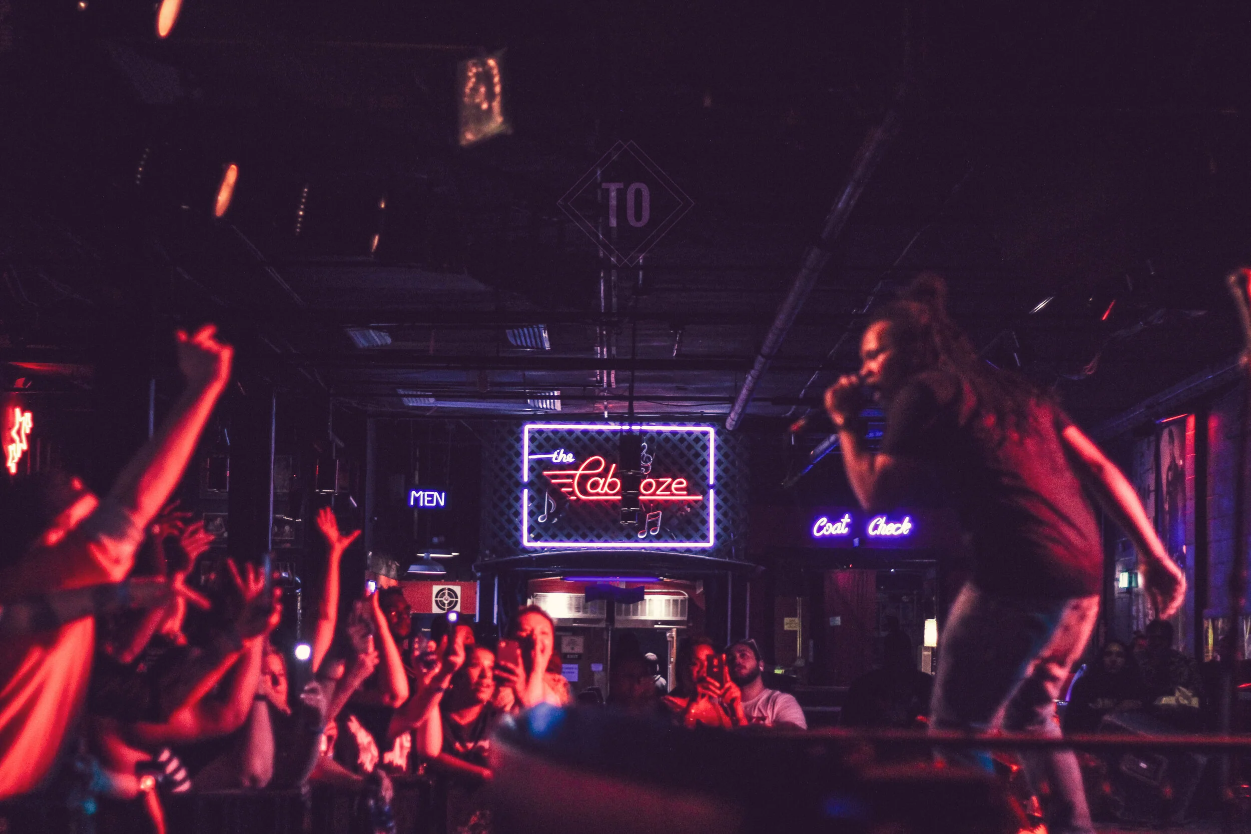 A singer performs on stage at a nightclub called 'the Cabareze' with neon signs and a crowd of people watching, some taking pictures, in a dark room with colorful lights.