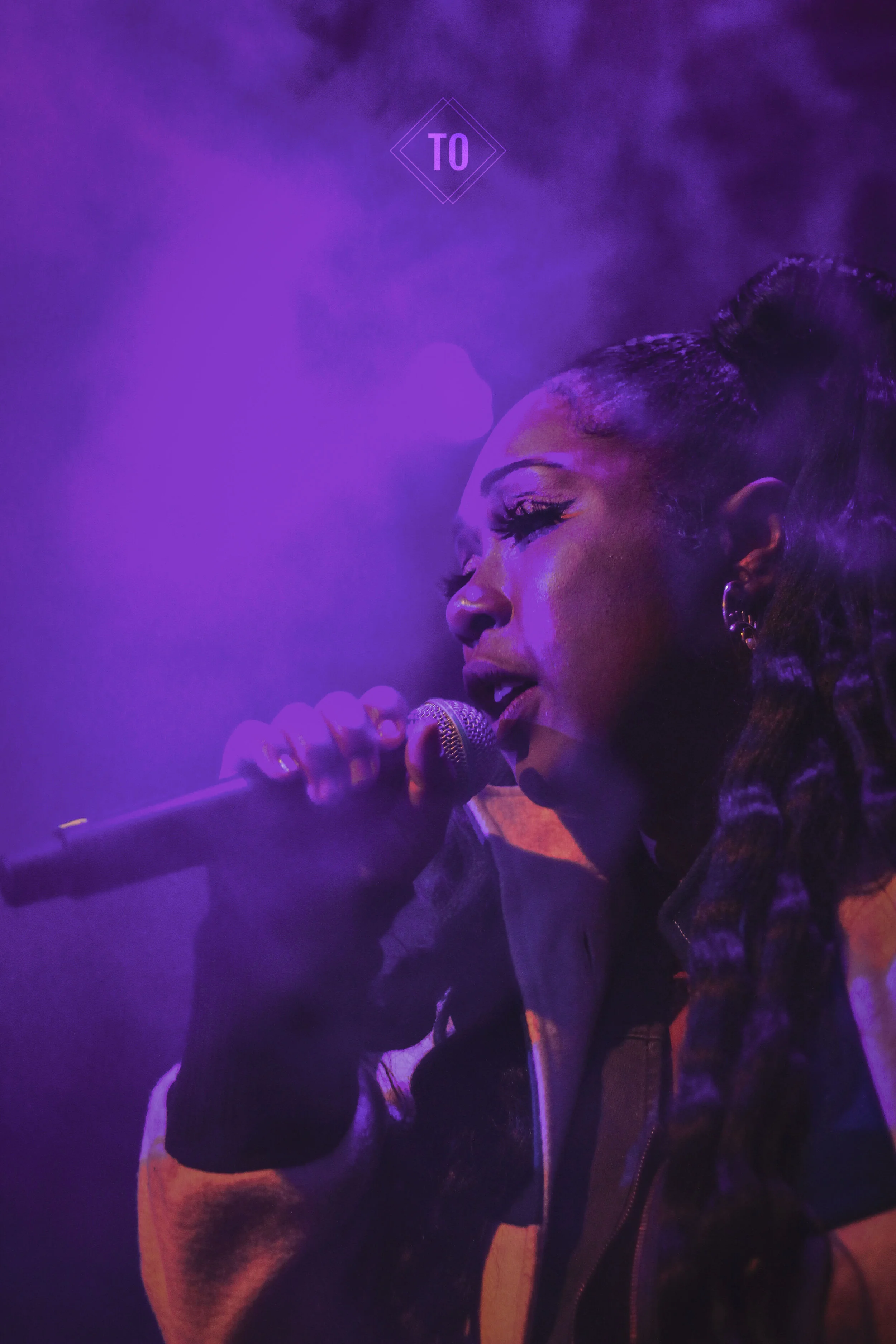 A woman singing into a microphone, illuminated by purple stage lighting.