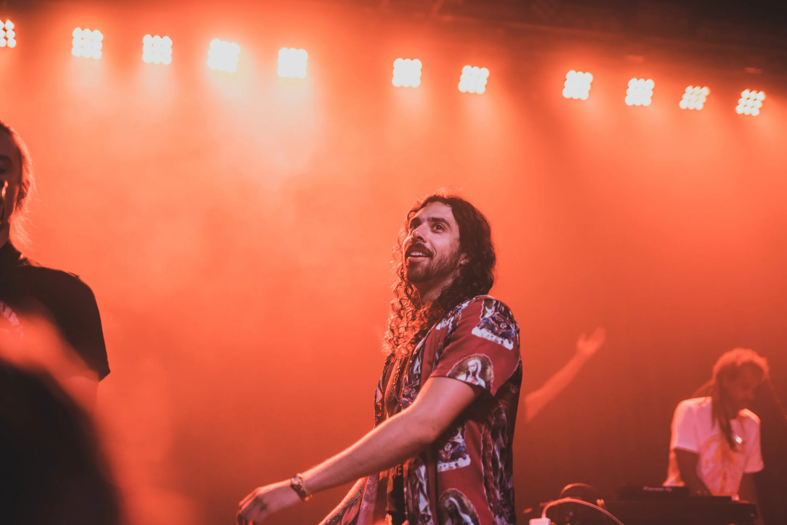 A musician with long curly hair and a beard performing on stage with orange lighting, accompanied by a DJ in the background.