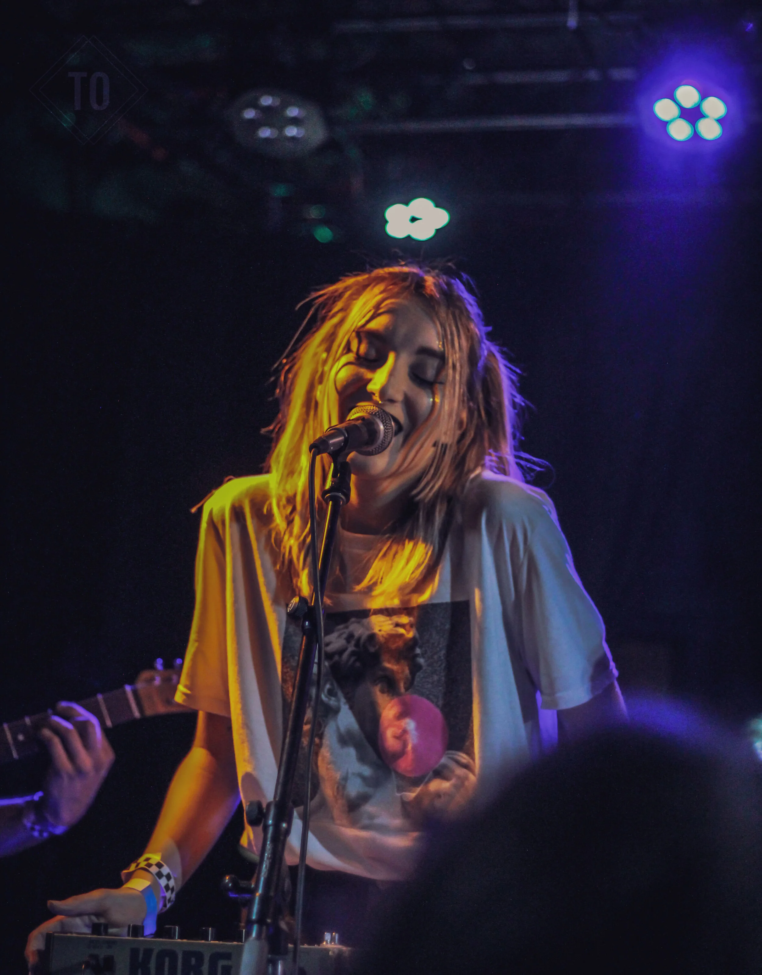 A woman with blonde dreadlocks singing into a microphone on stage, wearing a T-shirt with a graphic of a woman holding an apple, under colorful stage lights.