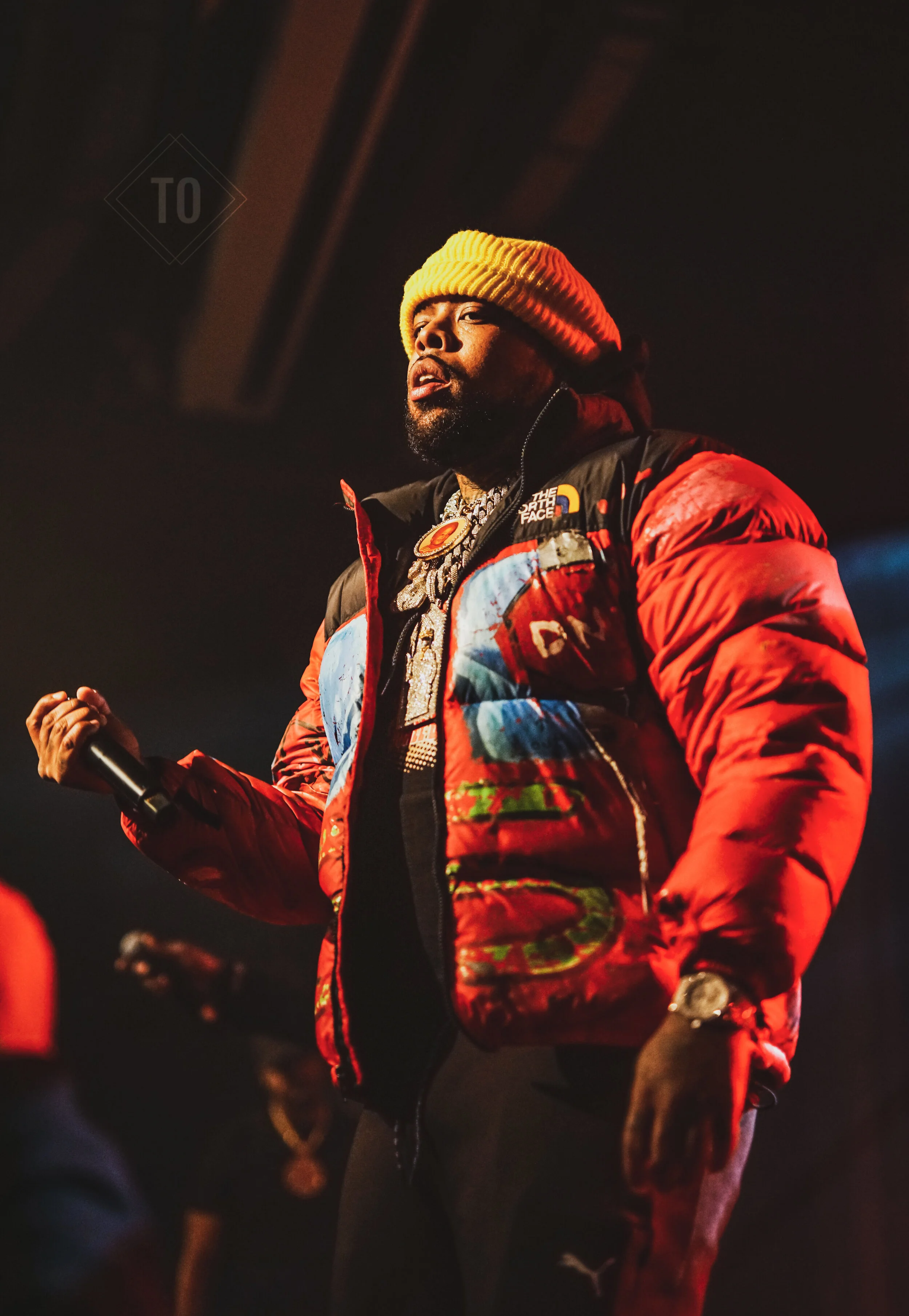 A man wearing a yellow beanie, black and red puffer jacket, and multiple chains, holding a microphone on stage during a performance.