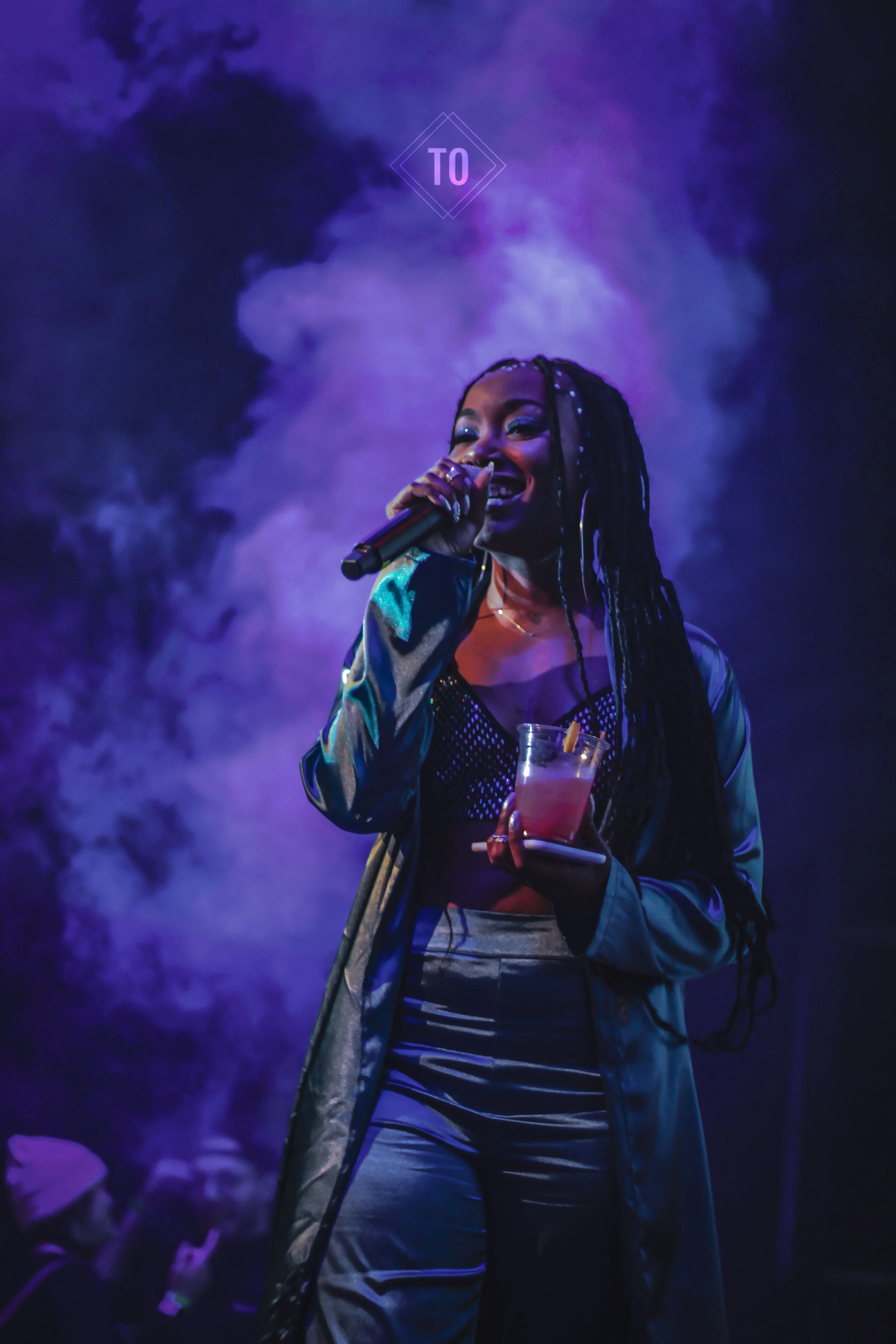 A woman with long braided hair holding a drink and a microphone, smiling on stage with purple smoke in the background.