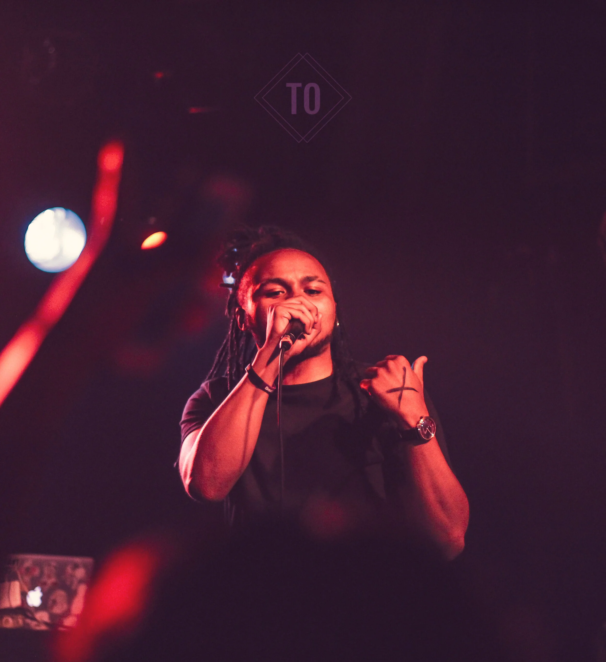 A person with dreadlocks singing into a microphone on stage, illuminated by pink and purple lighting.