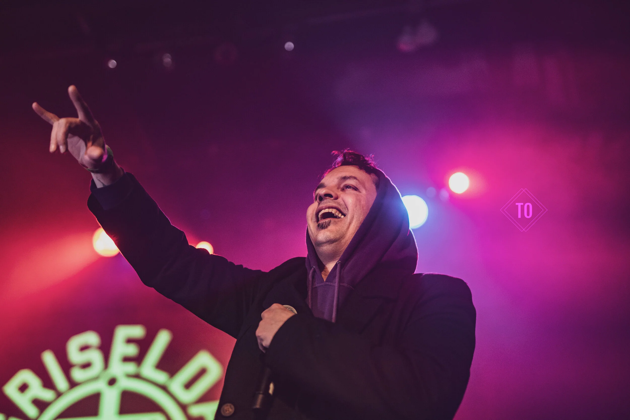 A man smiling and pointing upwards while holding a microphone at a concert with colorful stage lights in the background.