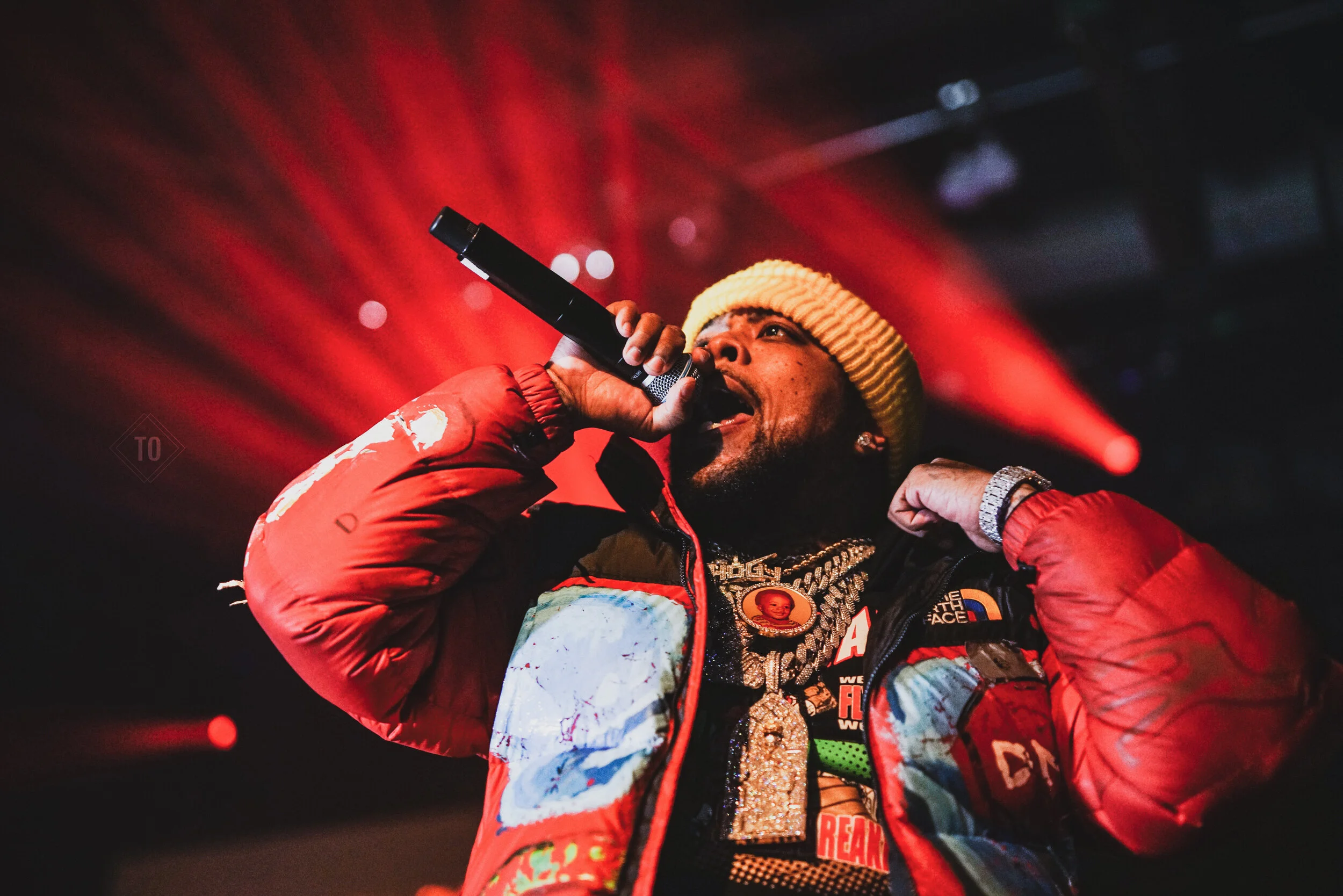 Performer singing into a microphone on stage, wearing a yellow knit cap, red puffy jacket, and jewelry, with red stage lighting in the background.