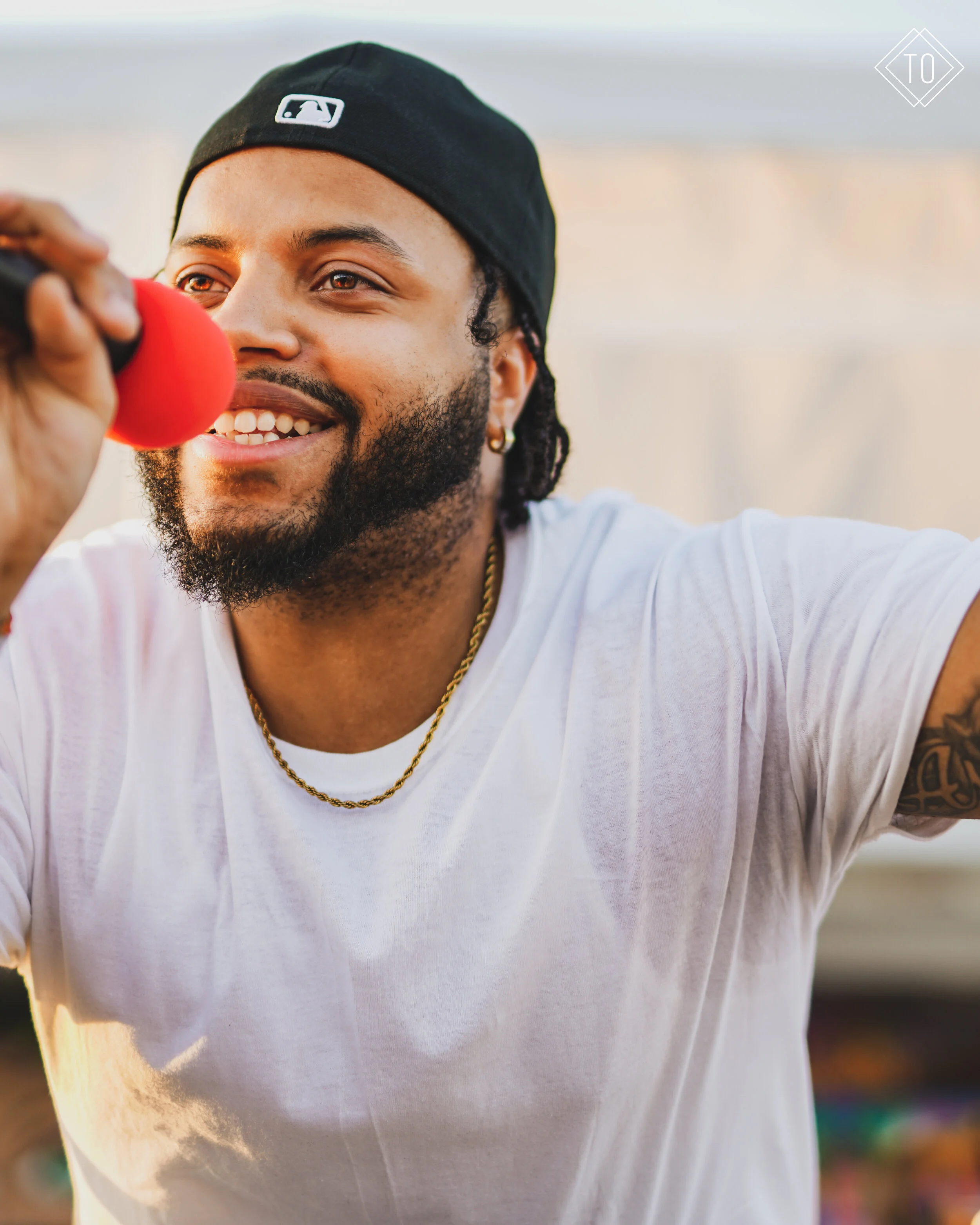 A smiling man with a beard and earrings, wearing a black baseball cap and white t-shirt, holds a red microphone.