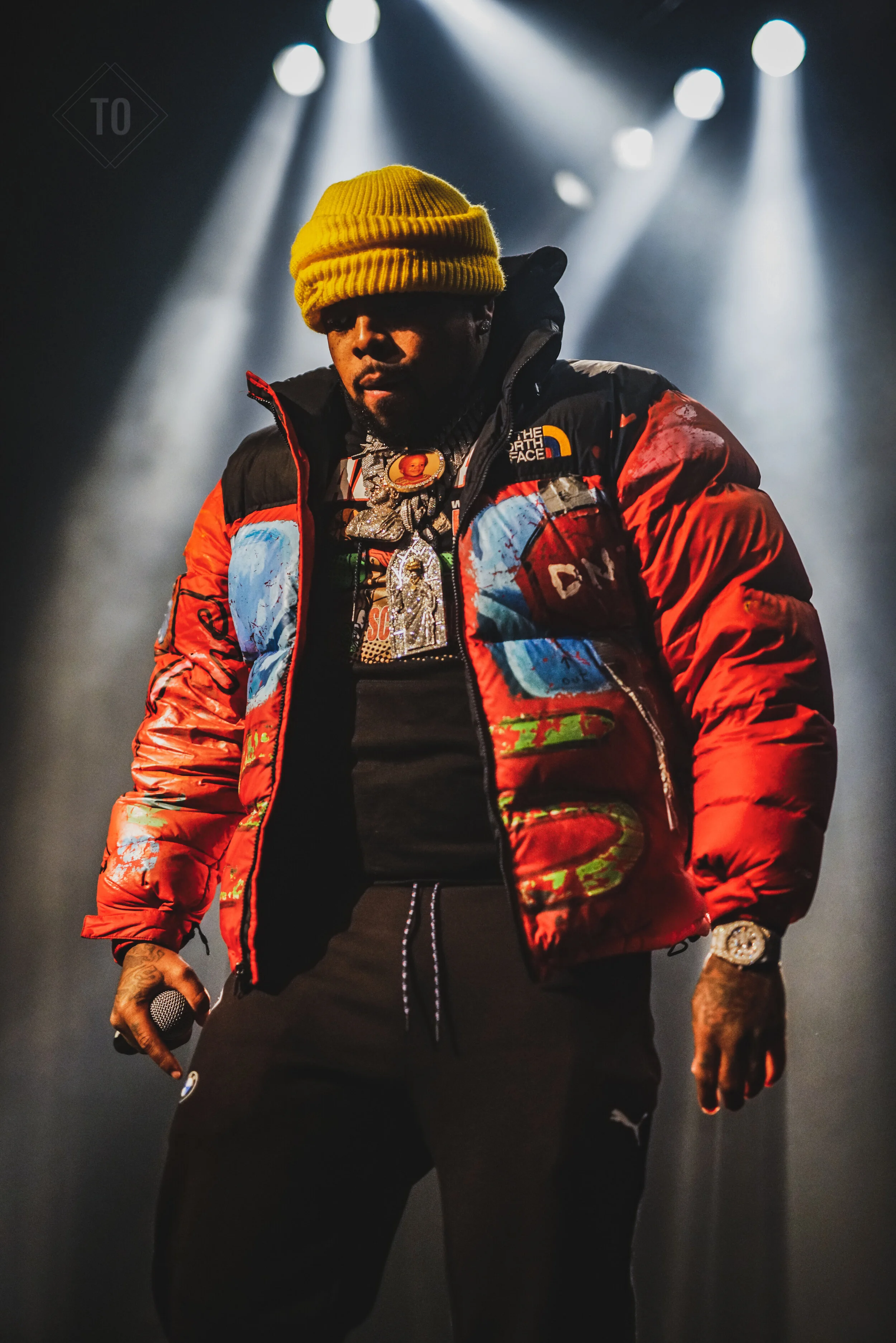 A male performer wearing a yellow beanie, a multicolored North Face puffer jacket, black pants, multiple chains, a watch, and holding a microphone on stage with spotlight beams overhead.