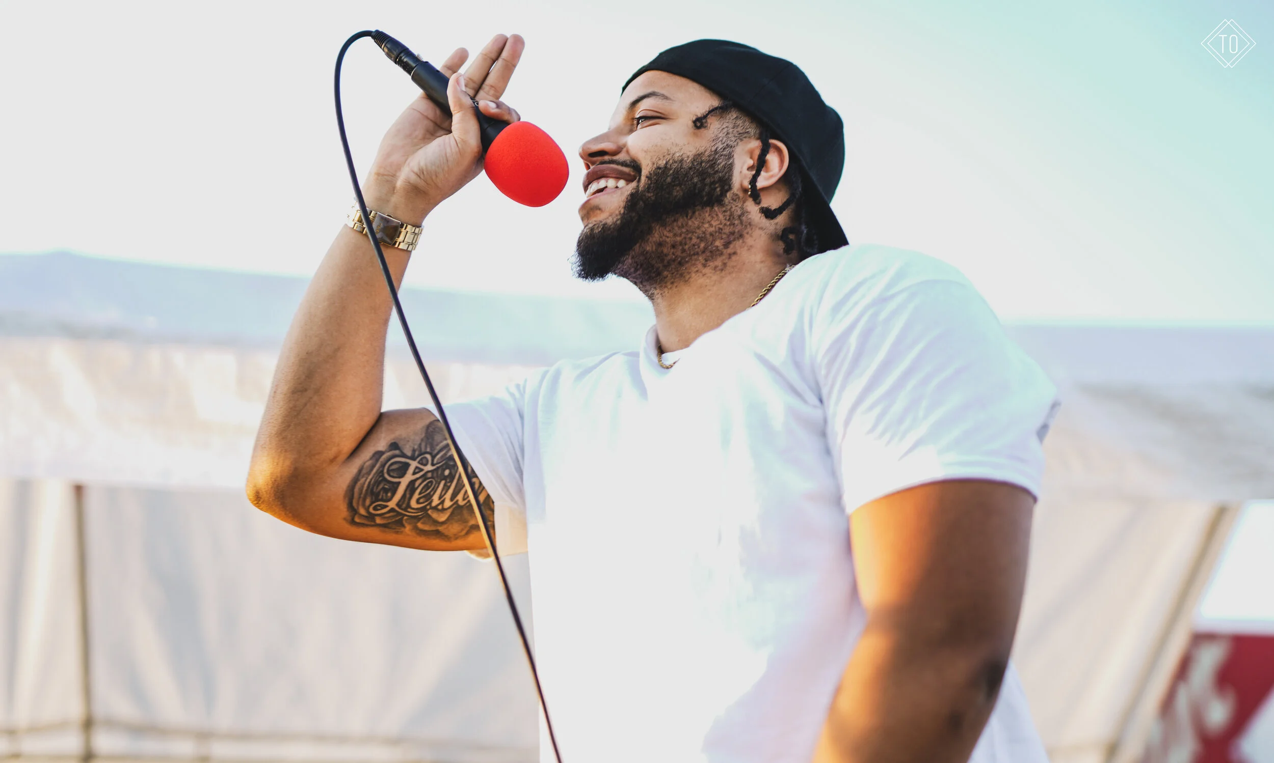 Man with a beard and tattoo wearing a white t-shirt, black cap, holding a microphone with a red windscreen, smiling outdoors.