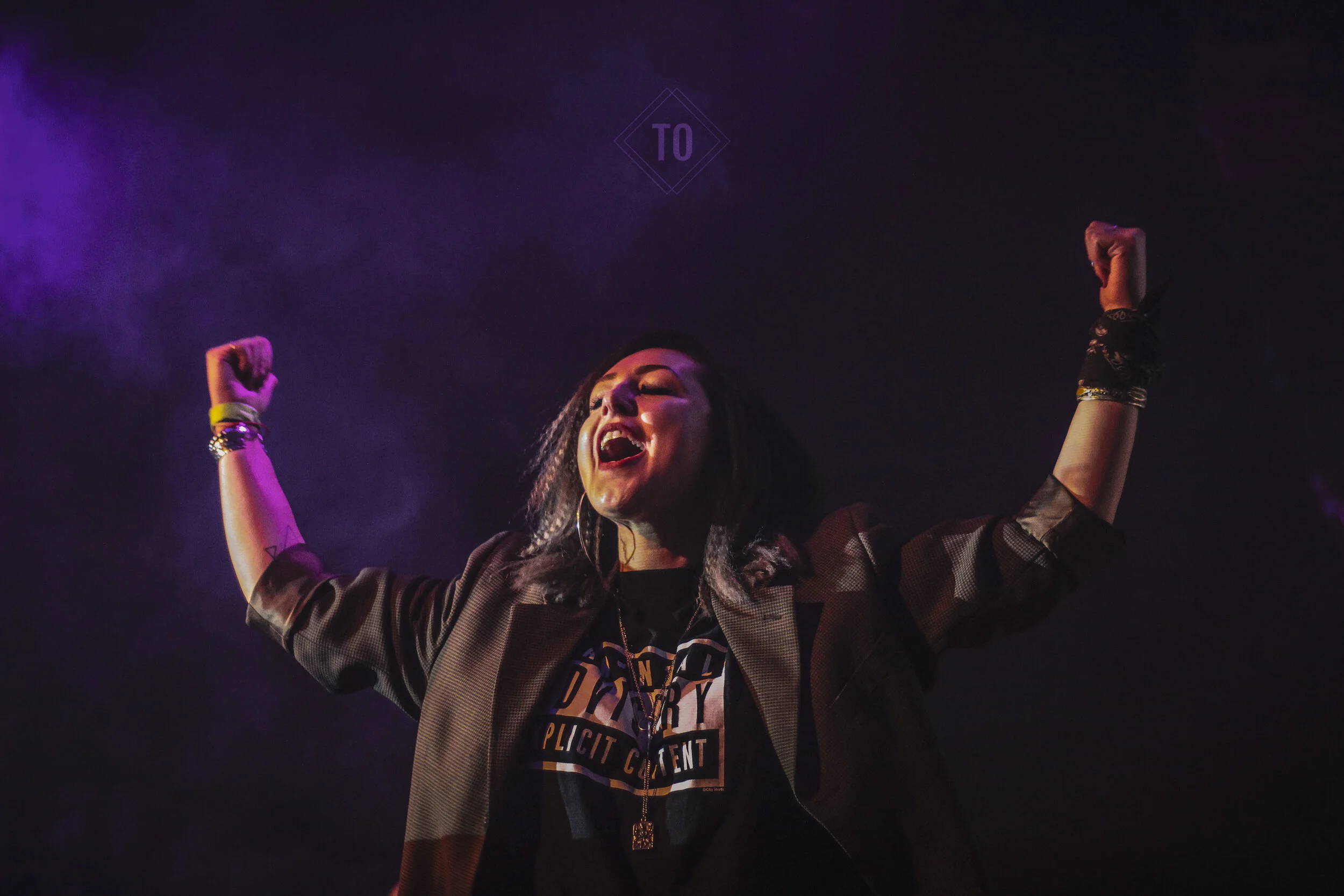 Woman with dark hair wearing jewelry and a dark T-shirt, raising her arms in victory or celebration with a wide smile, against a dark background with purple lighting.