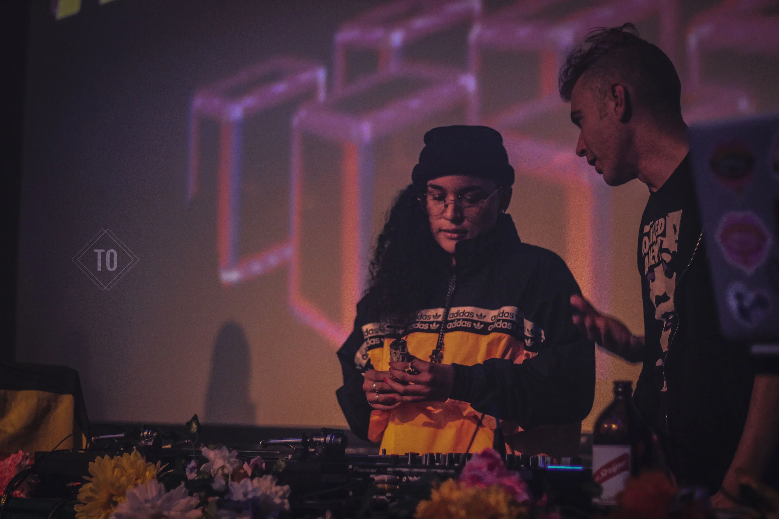 A woman and a man are standing behind a DJ setup with flowers on the table in front of them. The woman is wearing glasses, a black beanie, and a jacket. The man has a mohawk hairstyle and is wearing a black jacket with patches. They are looking at th