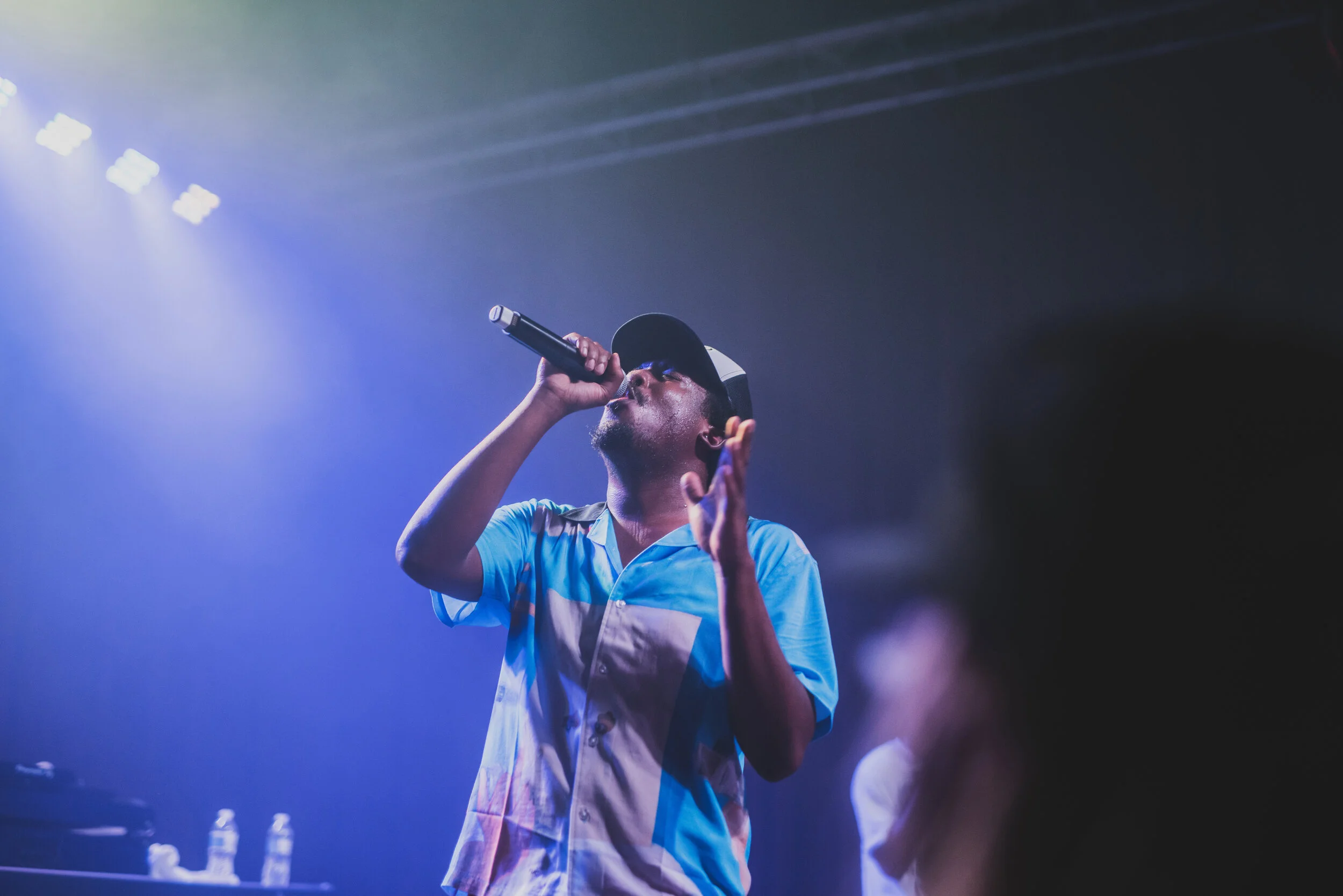 A male singer performing on stage, holding a microphone near his mouth, wearing a black cap and a short-sleeved shirt with blue, white, and pastel colors, with stage lights overhead.