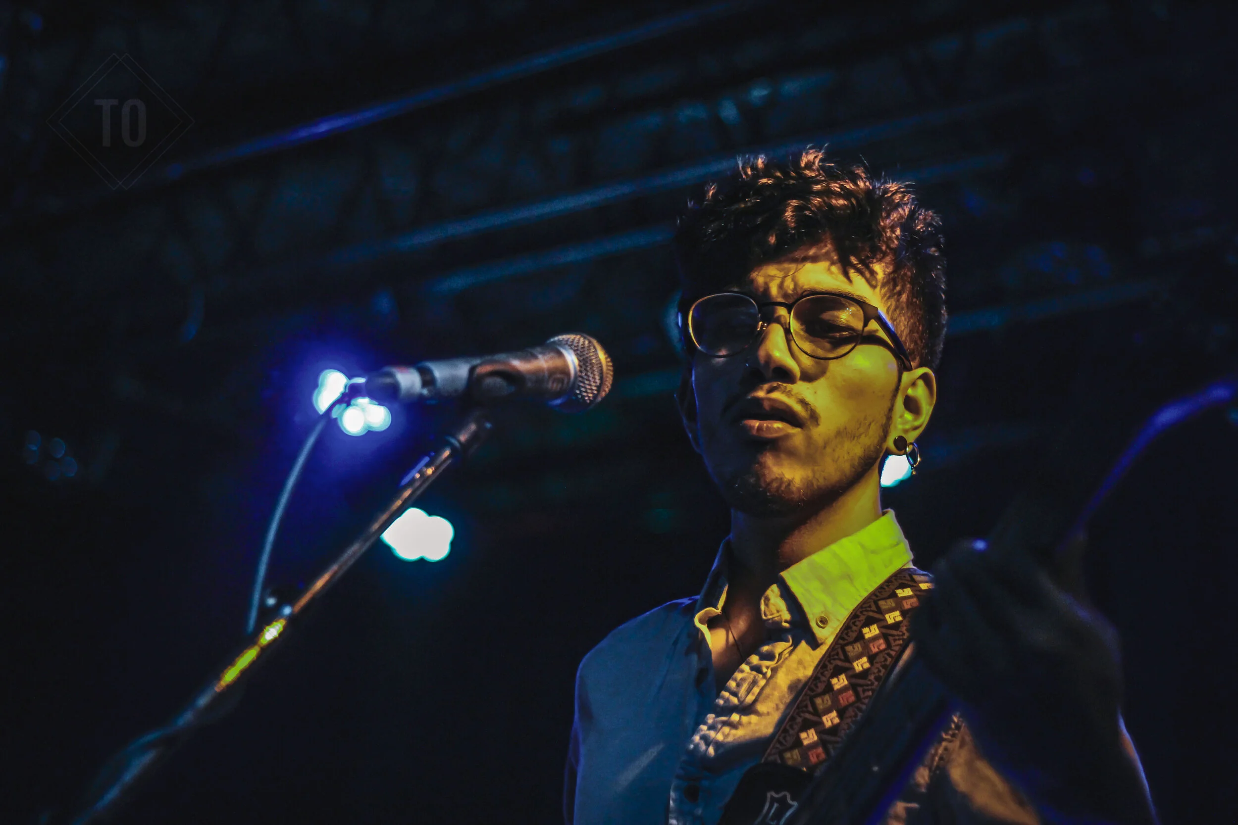 A young male musician with glasses and earrings performing on stage with a guitar, under blue and yellow stage lighting, near a microphone.