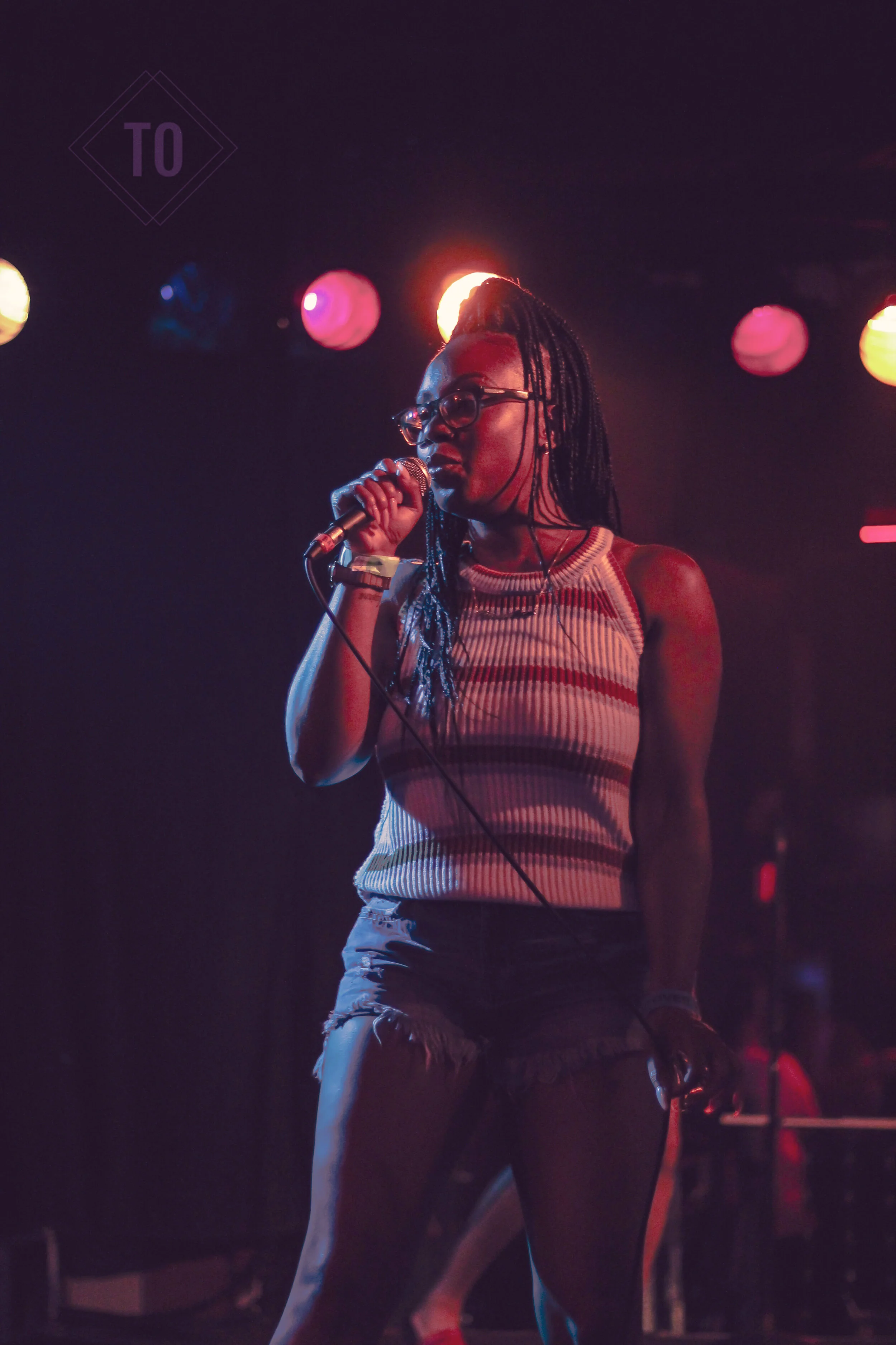A woman with glasses and long braided hair singing into a microphone on stage under colorful stage lights.