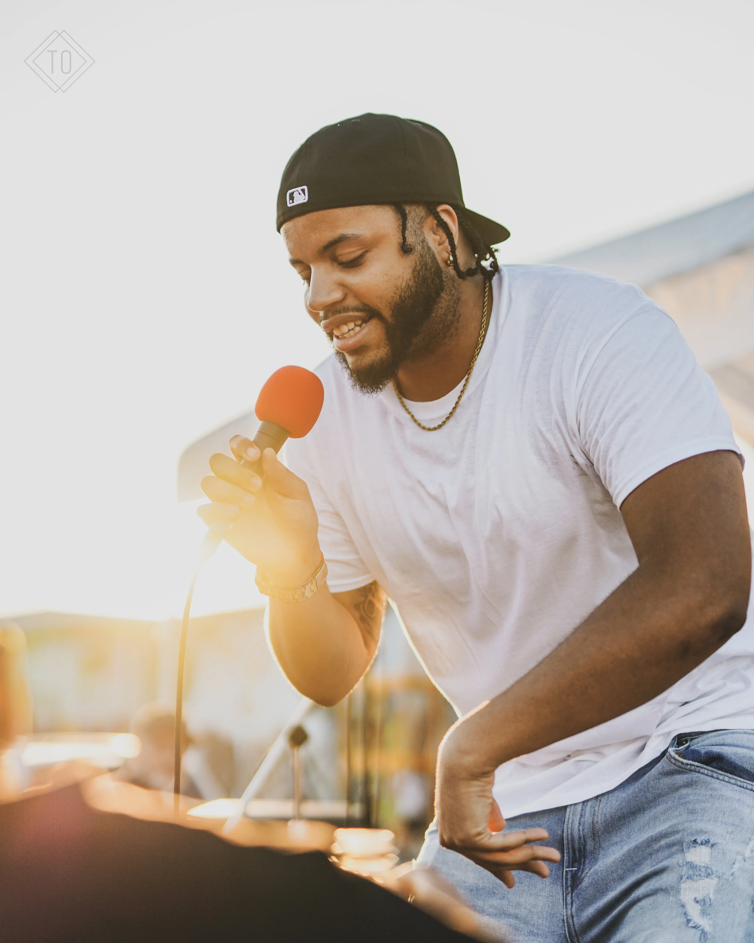 A man in a white t-shirt and black cap holding a red microphone and singing or speaking outdoors during sunset.